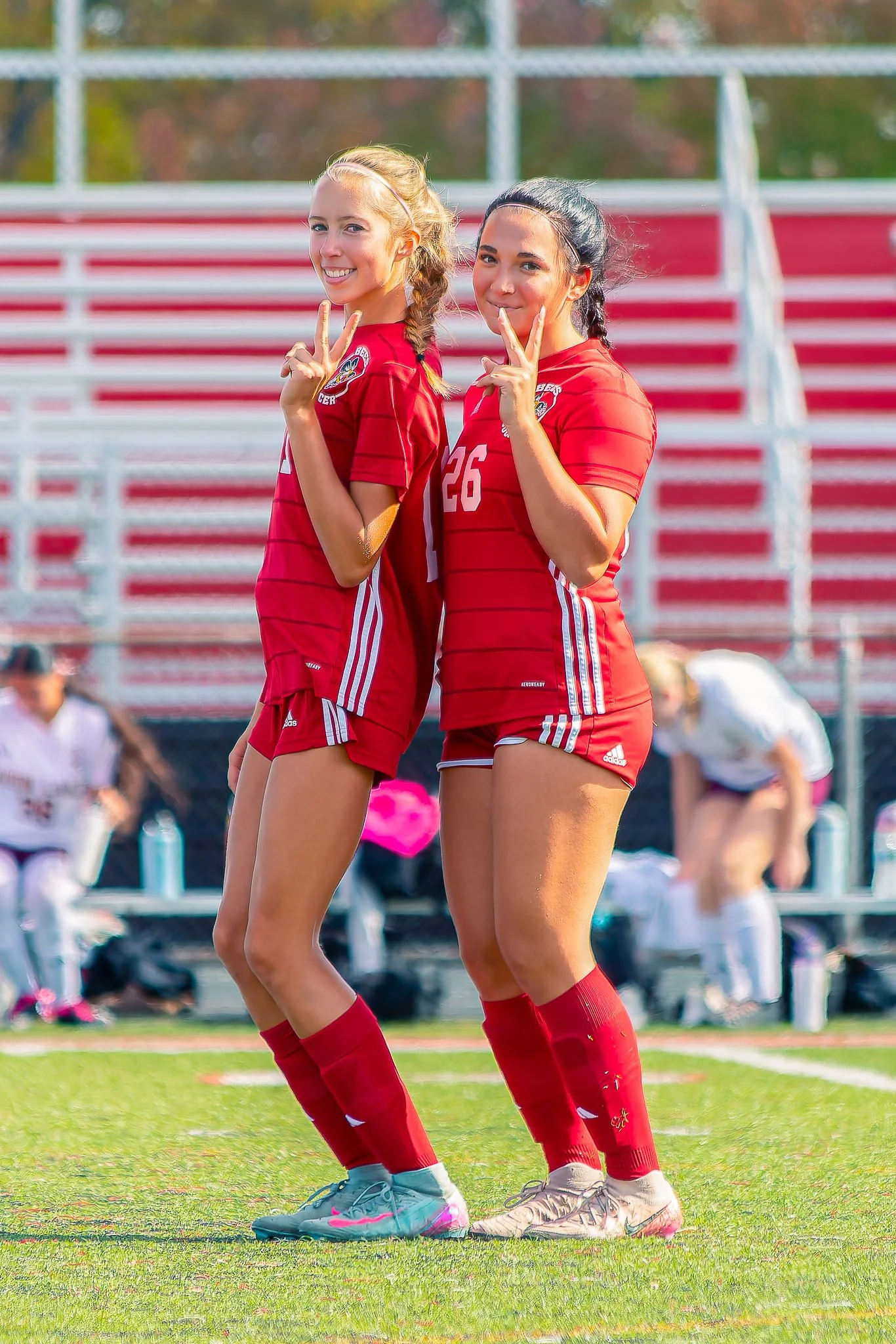 Two young female soccer players in red uniforms standing on the soccer field making peace signs with their hands and smiling at the camera.