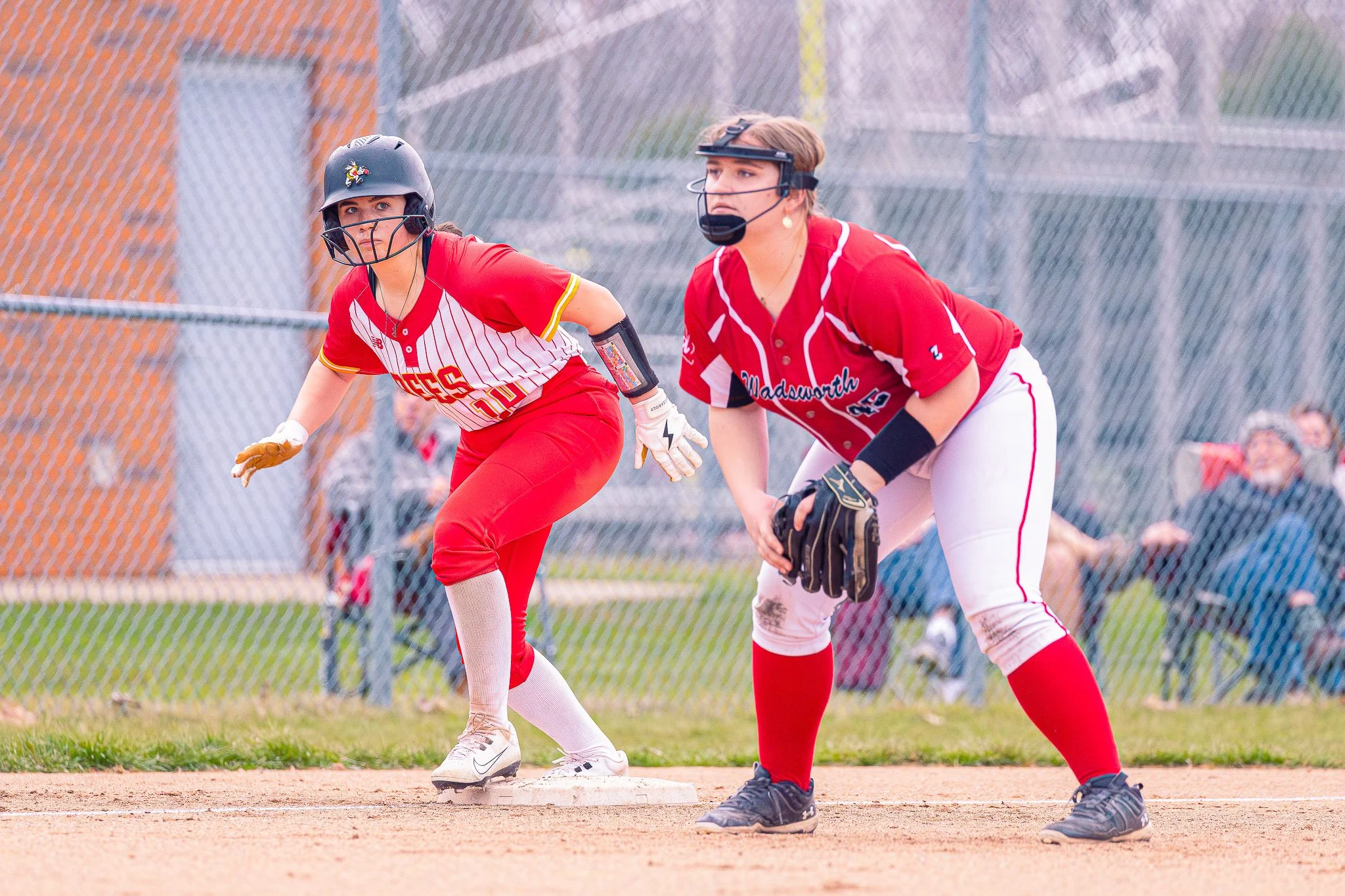 Two female softball players in red uniforms are positioned near a base on a softball field, preparing for a play. They wear helmets, gloves, and protective gear.