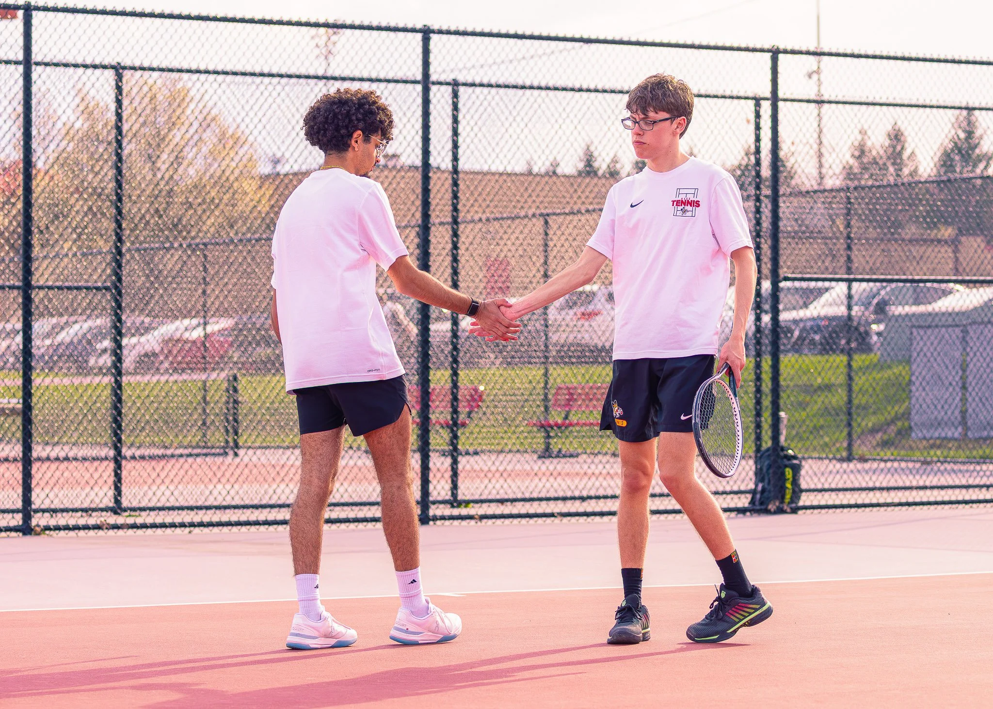 Two young male tennis players shaking hands on an outdoor tennis court at sunset.