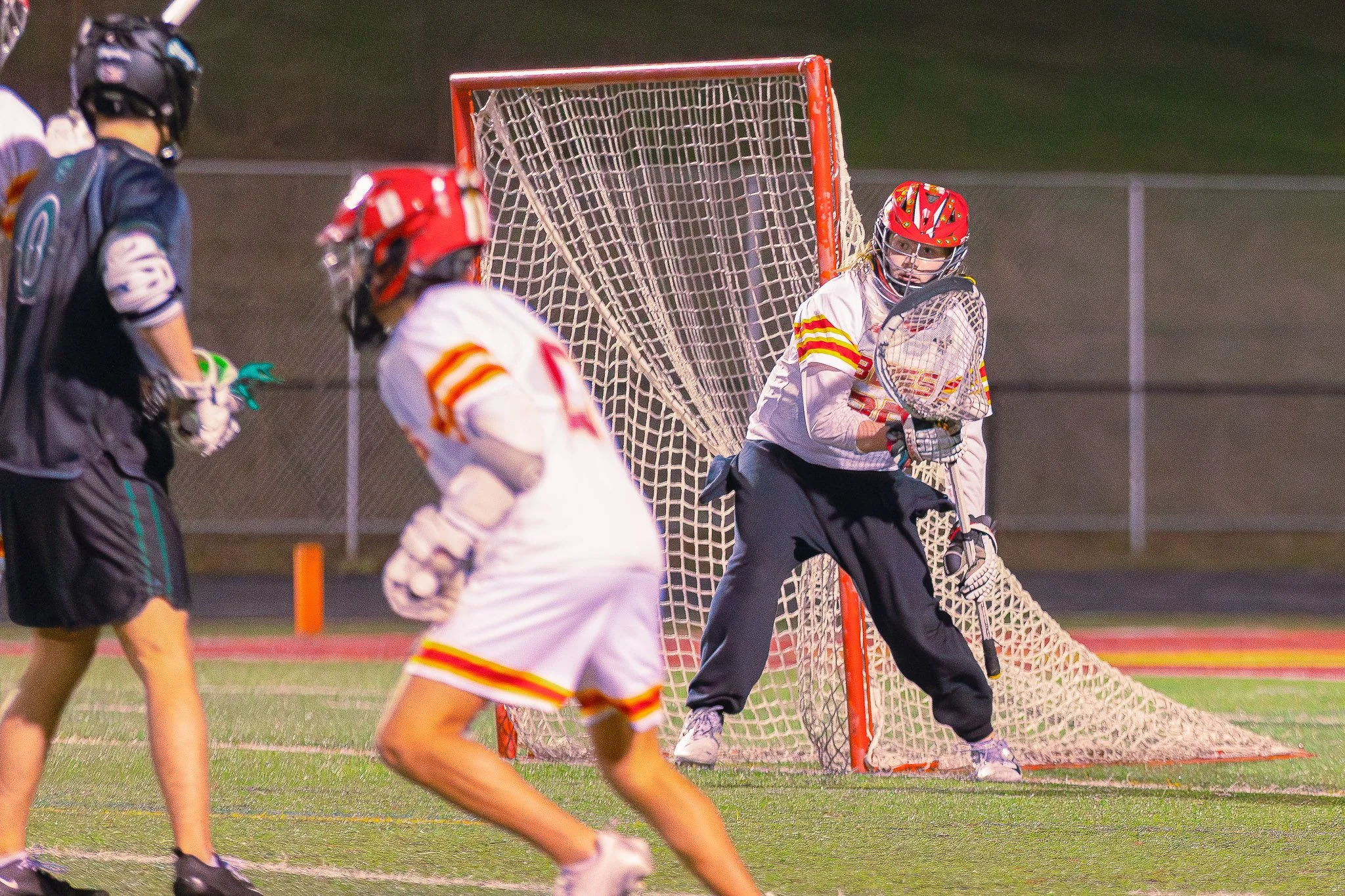 Lacrosse game at night with players near the goal, including a goalie in white with red and yellow striped accents, holding a lacrosse stick, defending the goal.