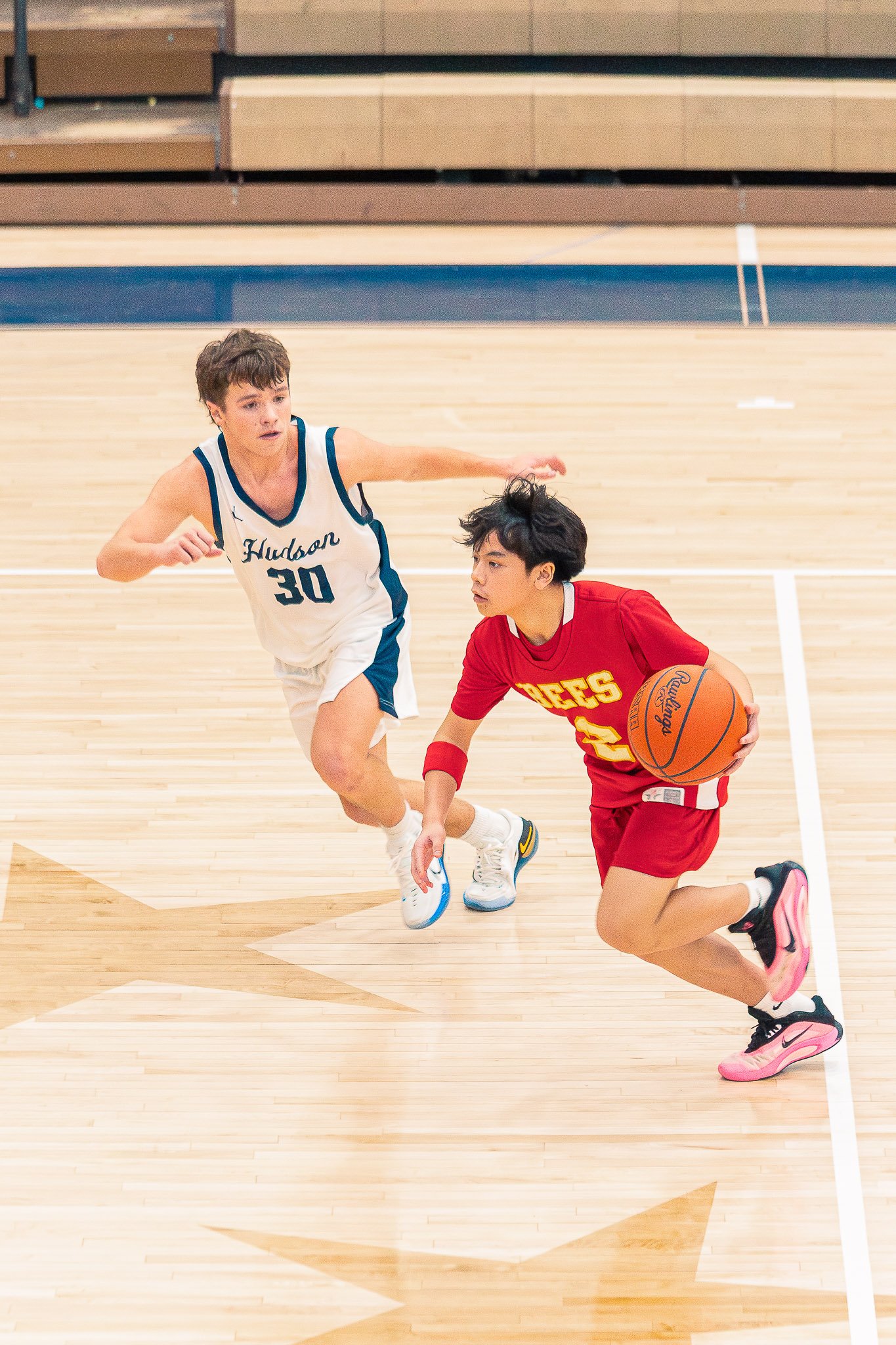 A young person in a red sports uniform dribbling a basketball while running on a wooden basketball court, with another young person in a white and blue sports uniform chasing behind.