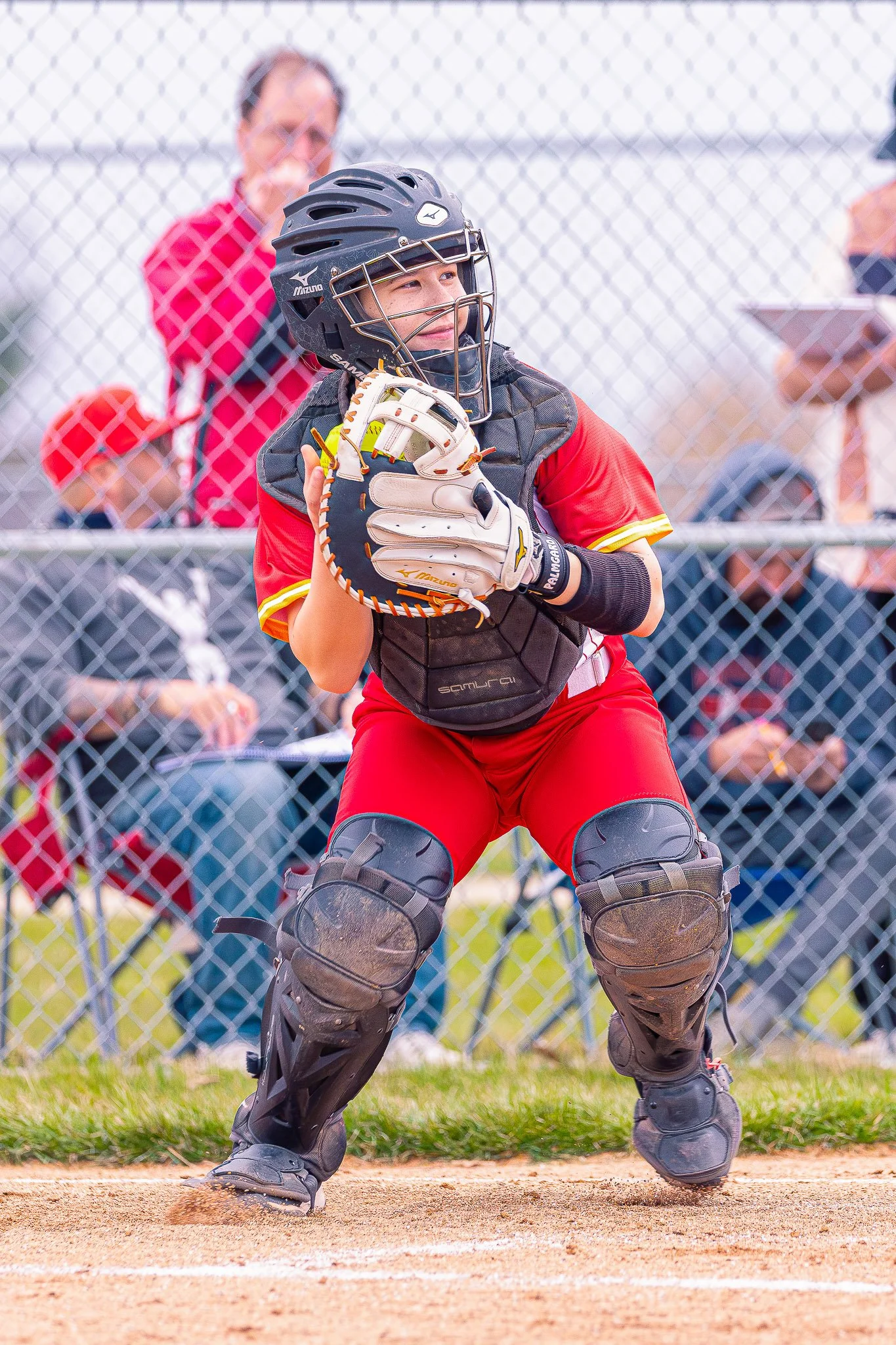 Softball player in catcher's gear crouching behind home plate, wearing a black helmet, black chest protector, and black leg guards, holding a baseball glove. Behind the player, a person in a red shirt is visible through a chain-link fence.