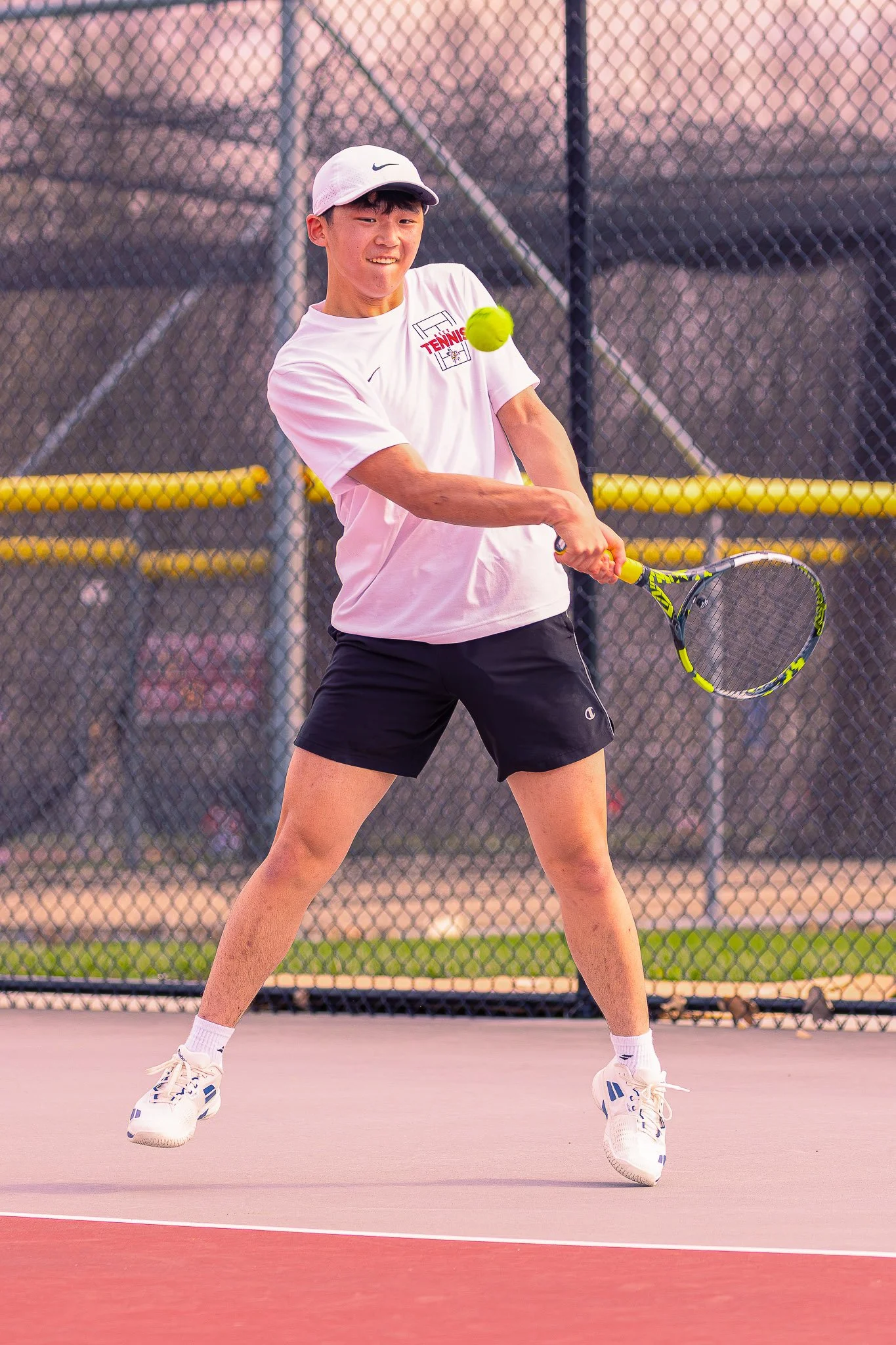 A young man wearing a white cap, white t-shirt, black shorts, and white sneakers is playing tennis on an outdoor court during sunset, with a tennis ball in mid-air near his racket.