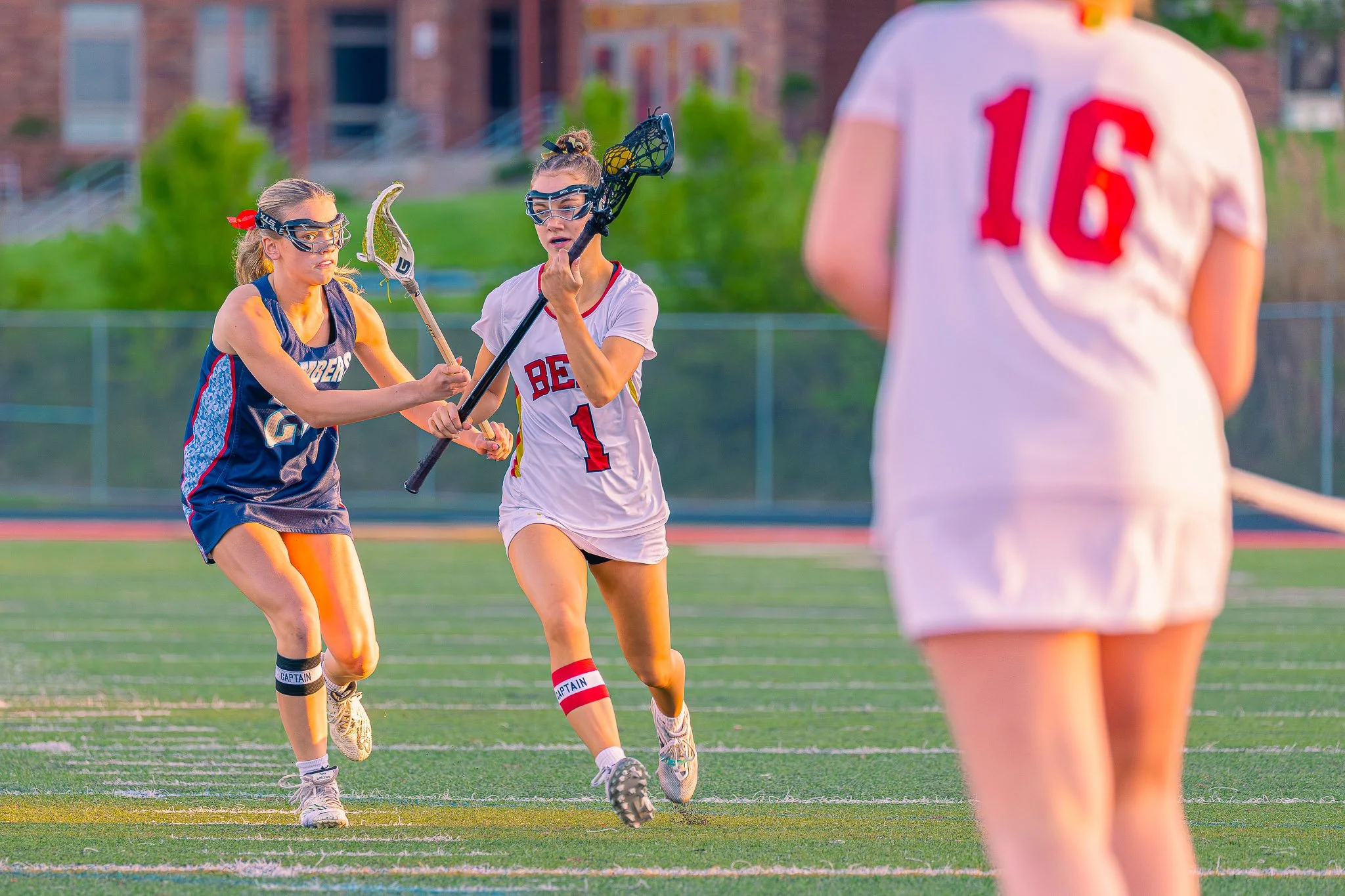Two young women playing lacrosse on a field, one wearing a white shirt with red accents and the other in a blue sports uniform, both with protective goggles, with a person in a white jersey with the number 16 in the foreground.