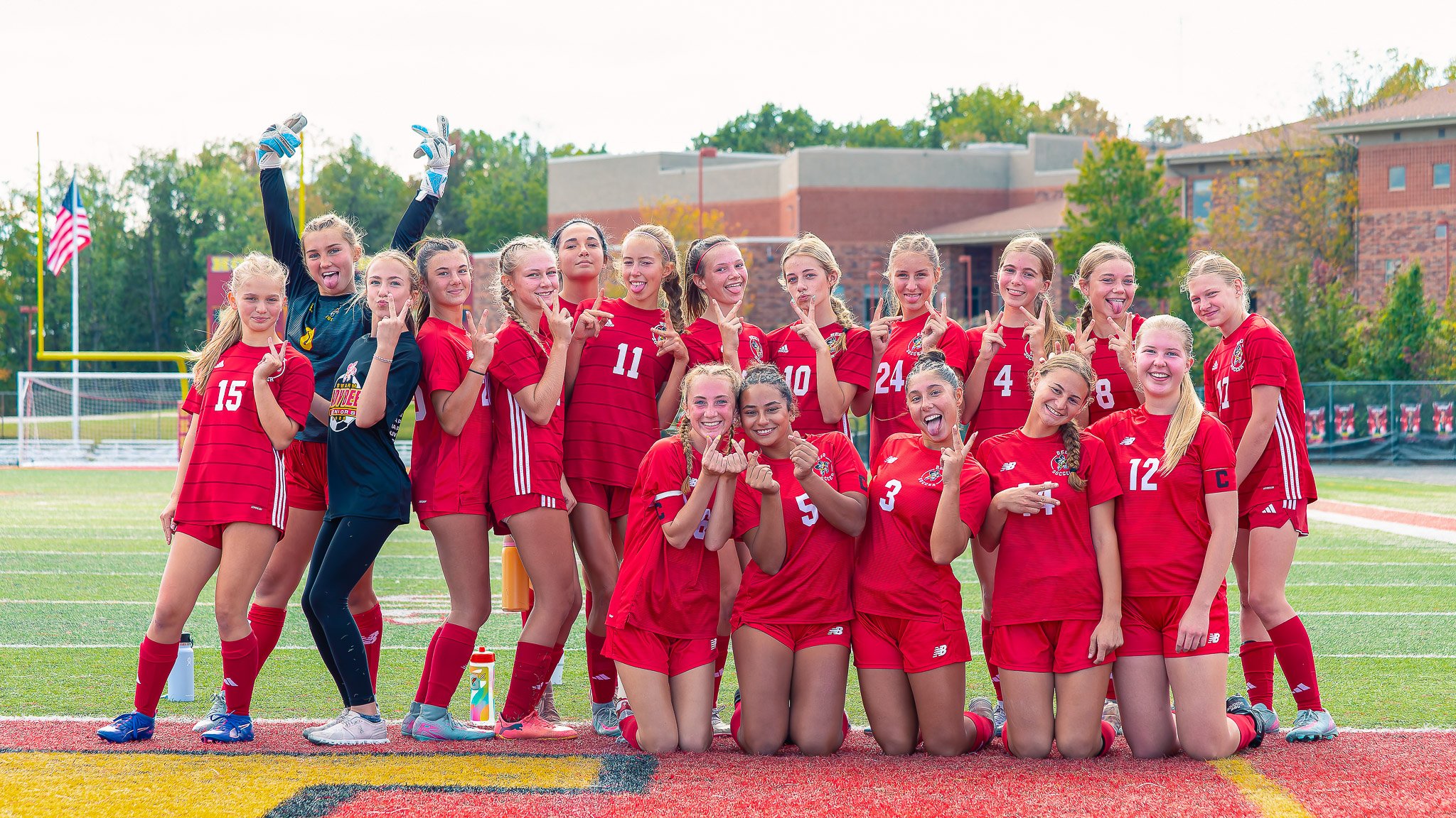 A group of female soccer players in red uniforms posing on a soccer field, smiling and making gestures, with goalposts and trees in the background.