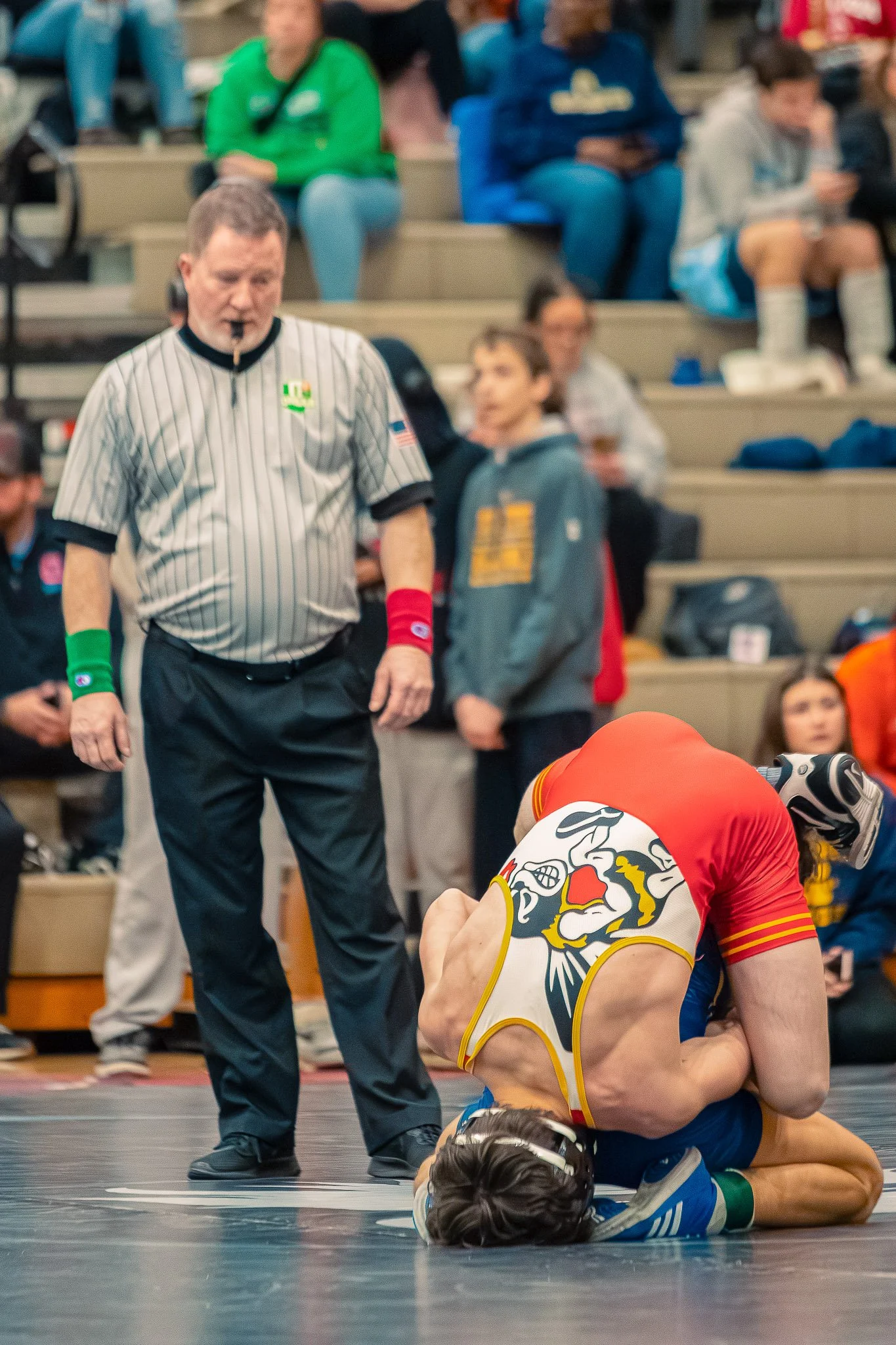 Wrestler in a yellow and blue singlet on the floor holding a wrestling opponent, with a referee and spectators in the background.