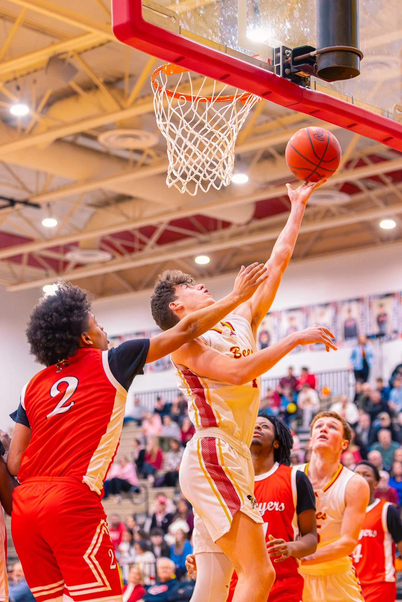 A basketball game in progress with players reaching for the ball near the hoop, occupied gymnasium with spectators in the background.