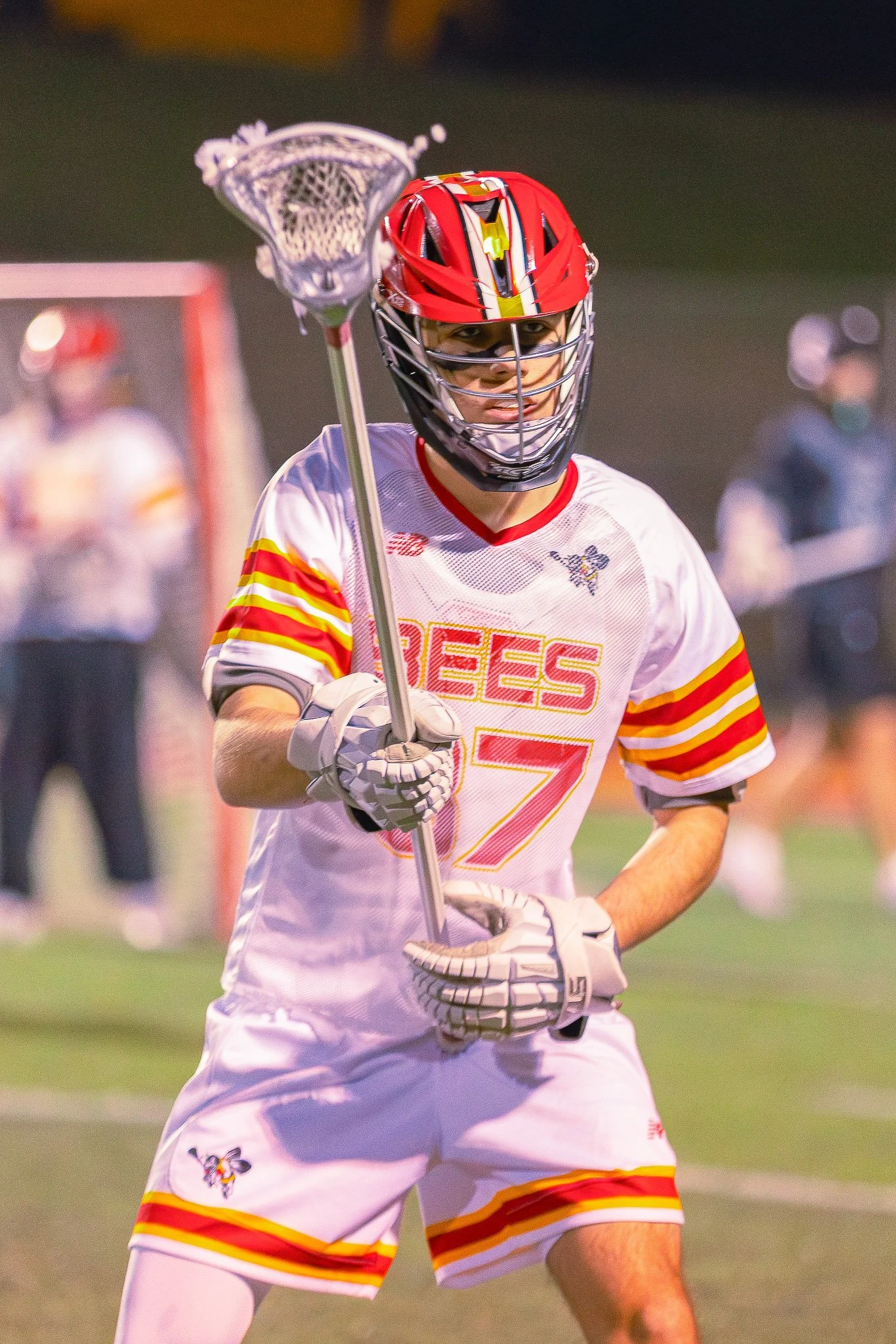 Lacrosse player wearing a red helmet and white jersey with red and yellow stripes, holding a lacrosse stick on a field during game.