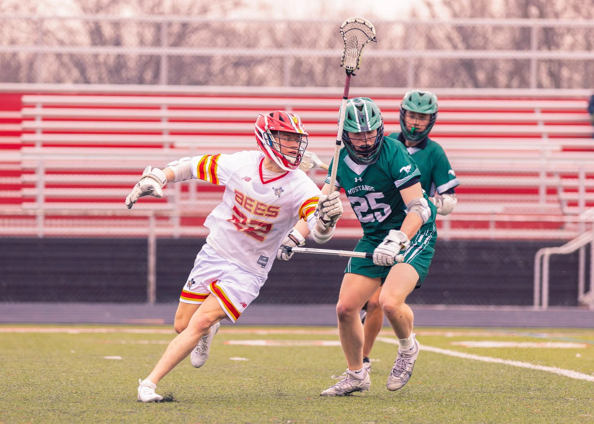 Lacrosse players competing on a field, one in a white and red uniform and another in a green and black uniform, wearing helmets and protective gear.
