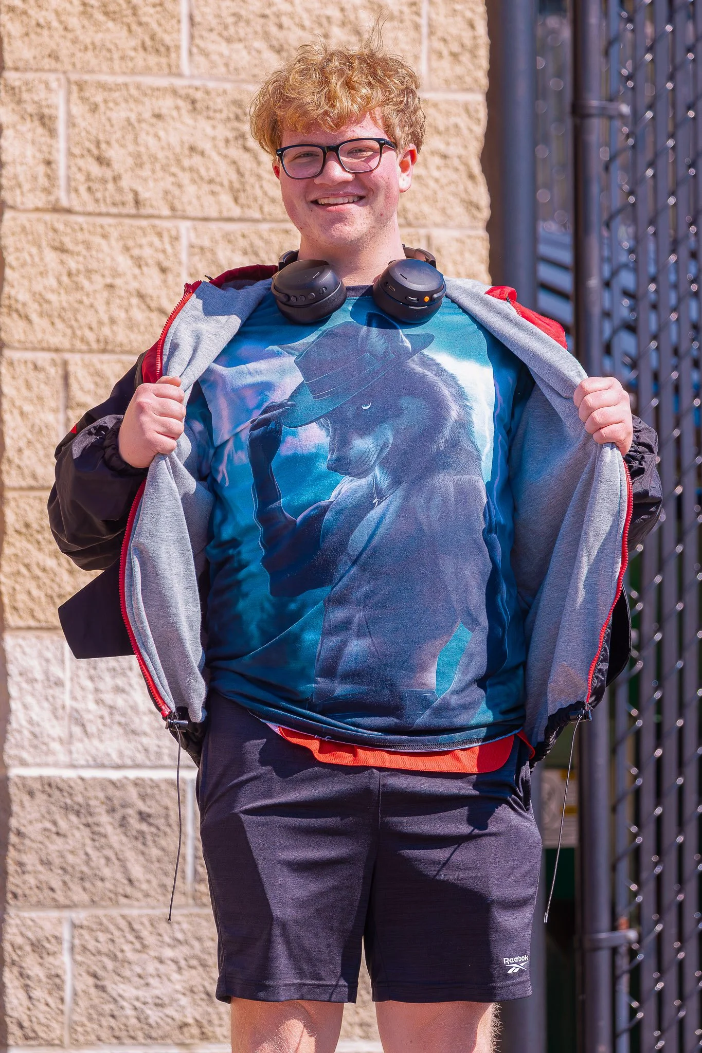 A young man with curly red hair and glasses standing outdoors, smiling, wearing a graphic T-shirt, shorts, and a jacket draped over his shoulders, with headphones around his neck.
