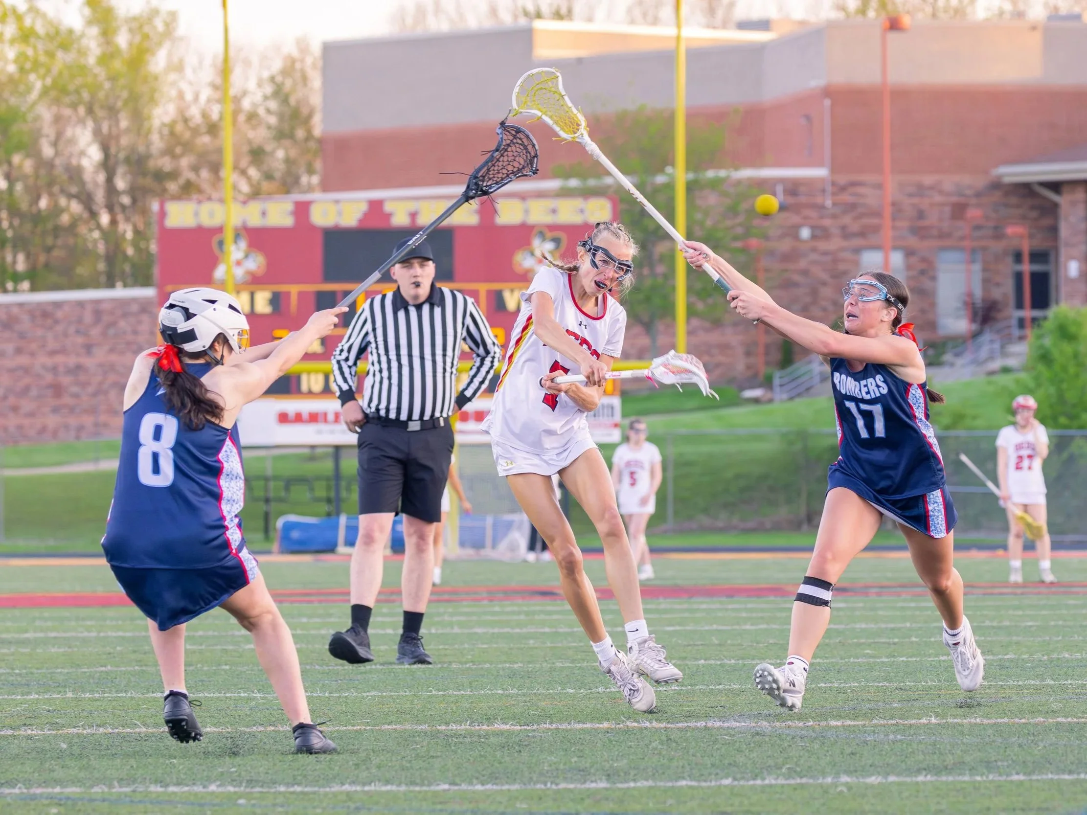 Women playing lacrosse on a field with a scoreboard and yellow goalposts in the background.