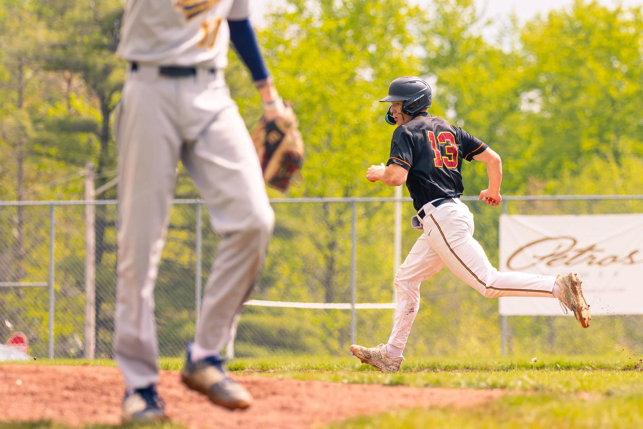 A young baseball player with a black helmet and black jersey numbered 13 running on the field. An opposing player in white uniform with gray pants, a yellow and black glove, and a backpack is in the foreground. The background shows green trees and a 