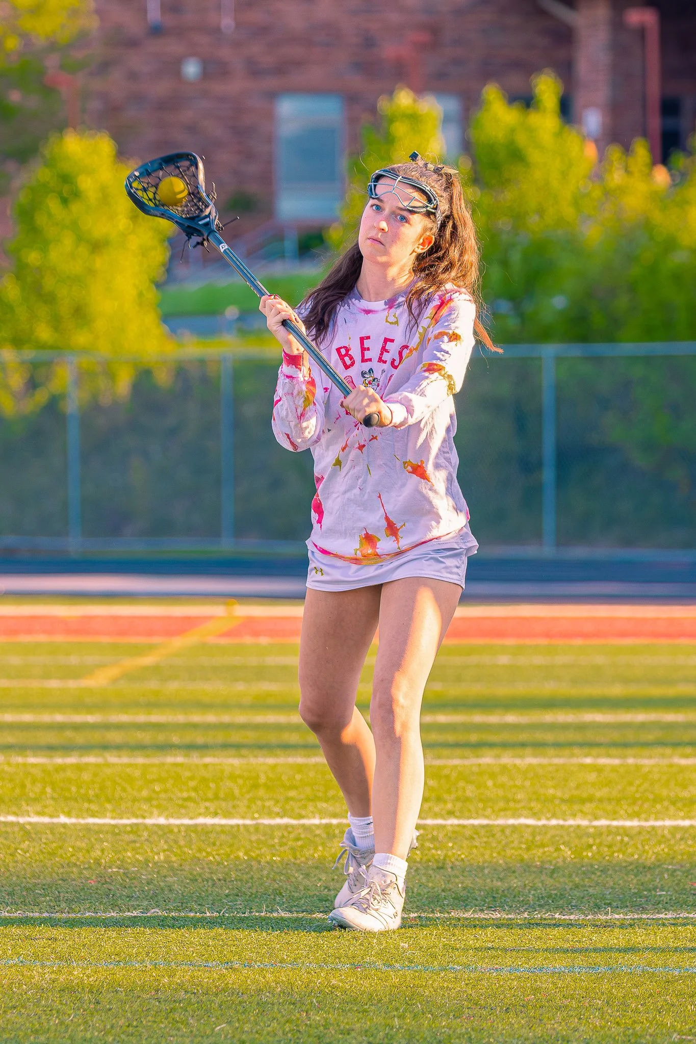 A young woman playing lacrosse on a field with trees and a building in the background, holding a lacrosse stick with a yellow ball in the net.