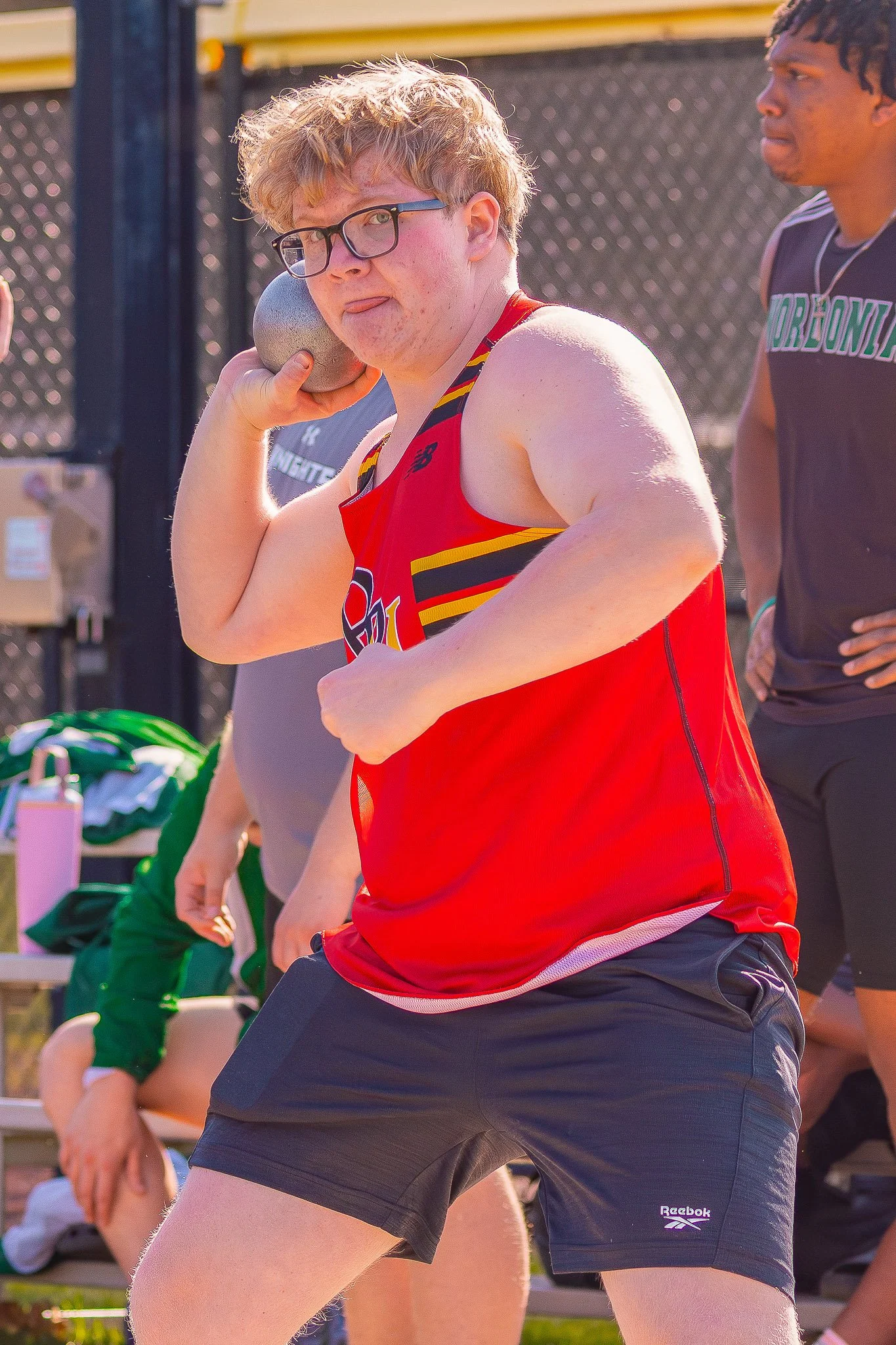 A group of people participating in a shot put competition outdoors, with one person holding a shot put near his neck and others standing around.