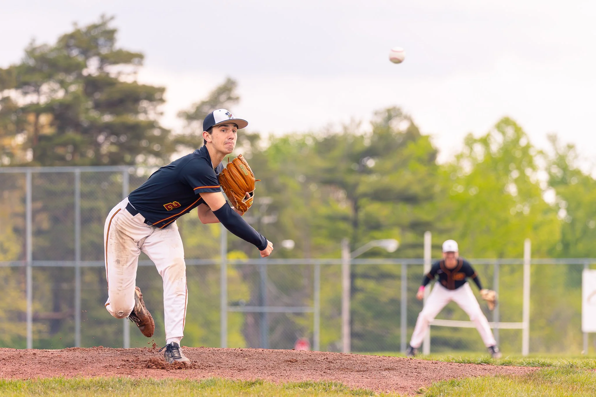 A baseball player in a black jersey and white pants is pitching on a baseball field, with a teammate in the background and trees in the distance.