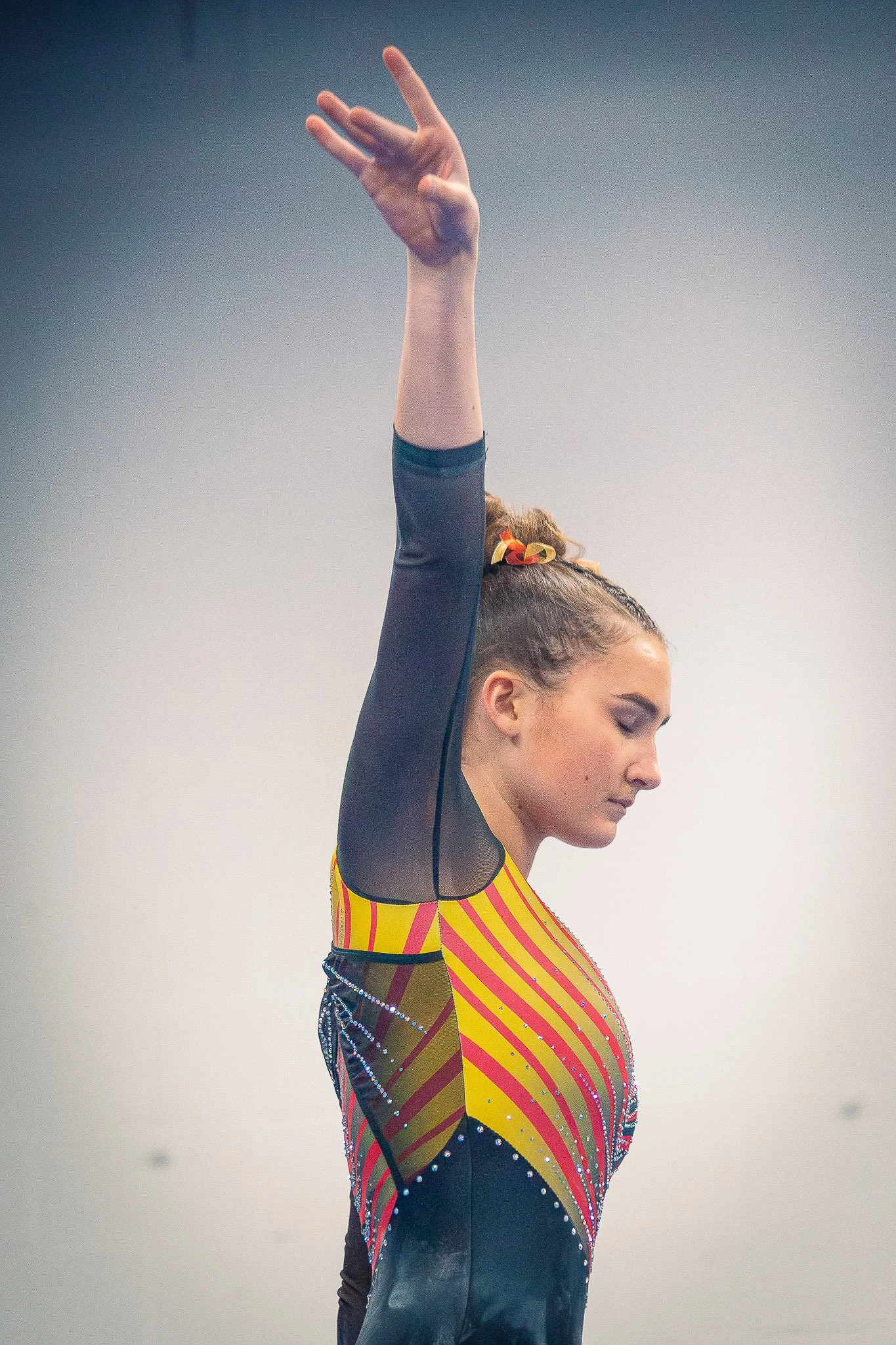 Gymnast standing with eyes closed, one arm raised with fingers extended, wearing a colorful, striped leotard with rhinestone embellishments.