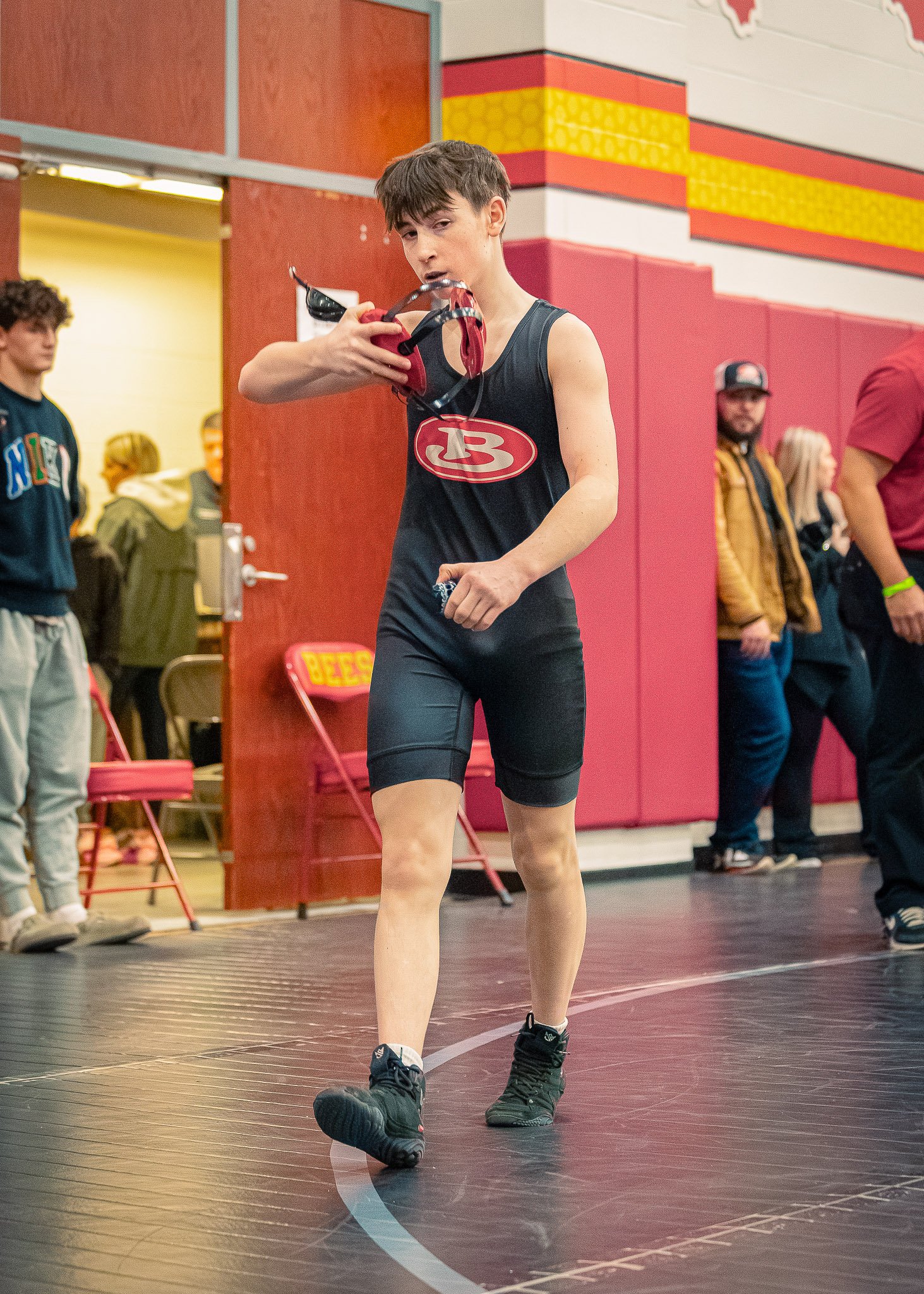 A young male athlete in a black sleeveless athletic outfit with a red and white logo, standing on a black indoor basketball court, holding goggles and looking at the camera. Behind him, other people are present, some standing, some sitting, with a re