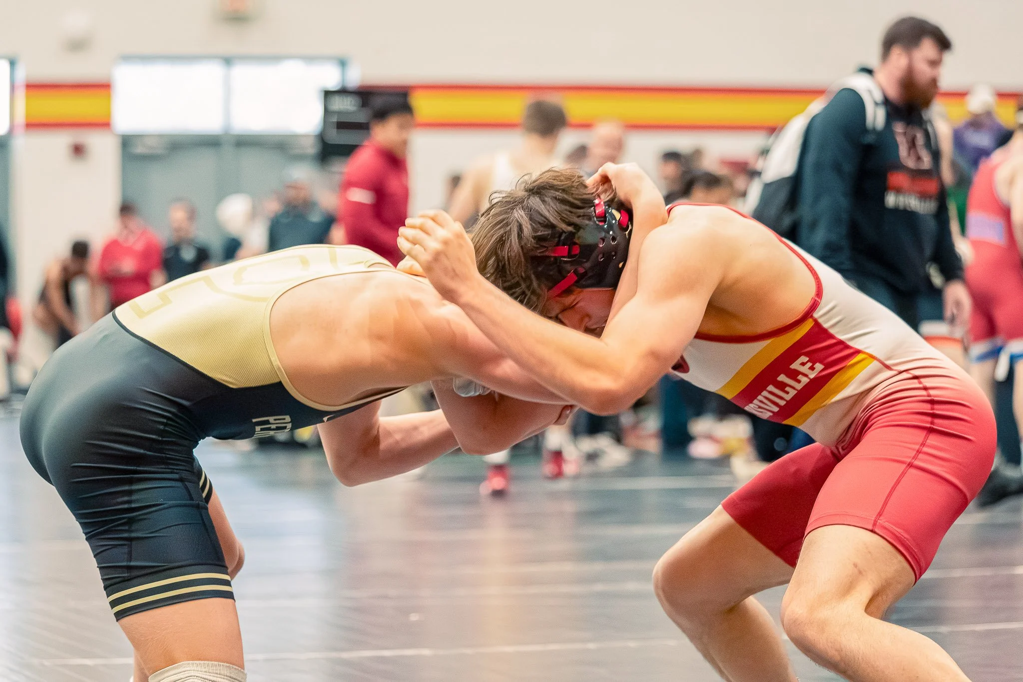 Two wrestlers wearing singlets and headgear engaged in a wrestling match, leaning into each other in a gymnasium with spectators in the background.