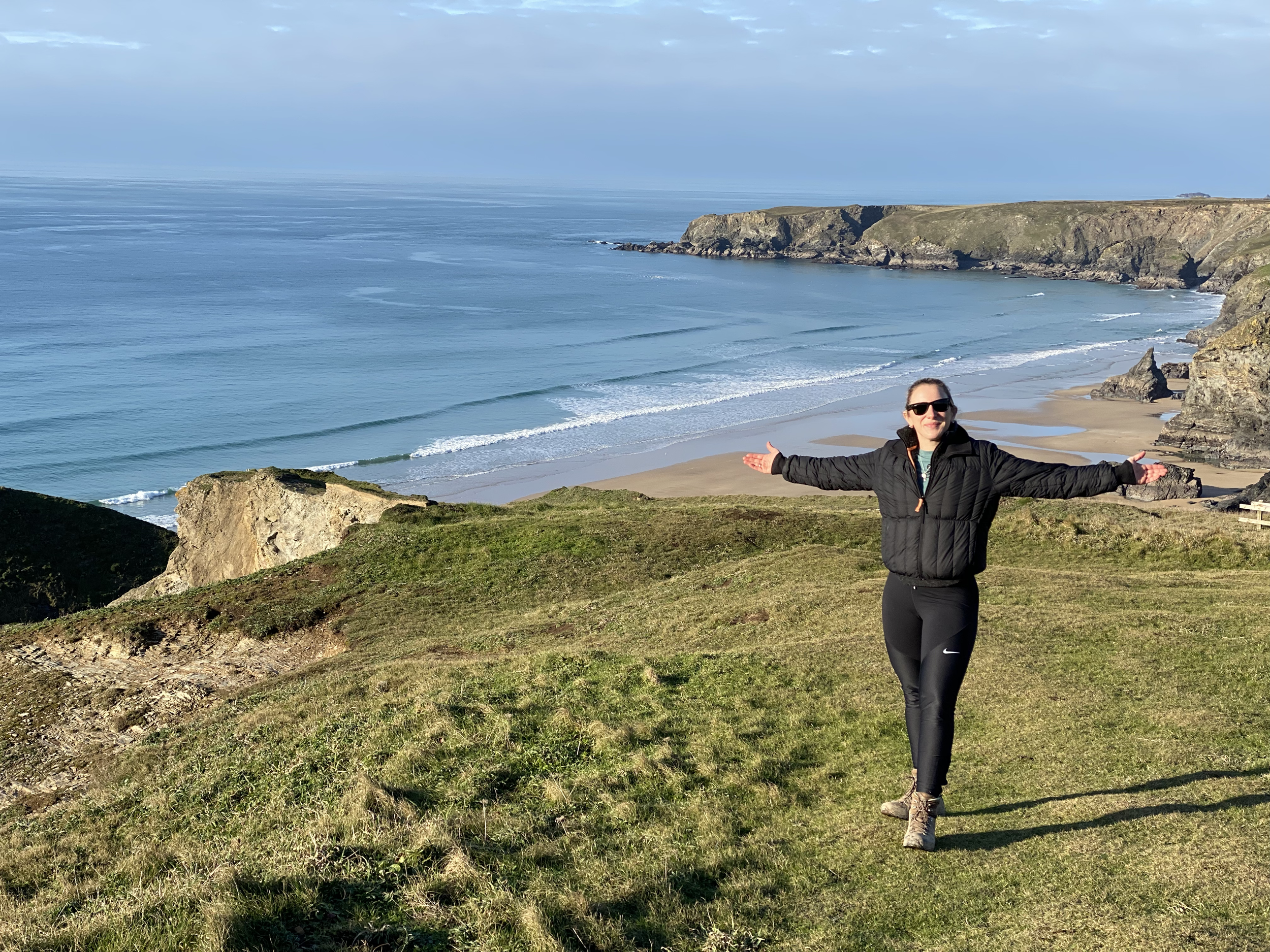 A woman standing on a grassy cliff edge with arms out wide, overlooking the ocean with waves and a distant cliff on a clear day.