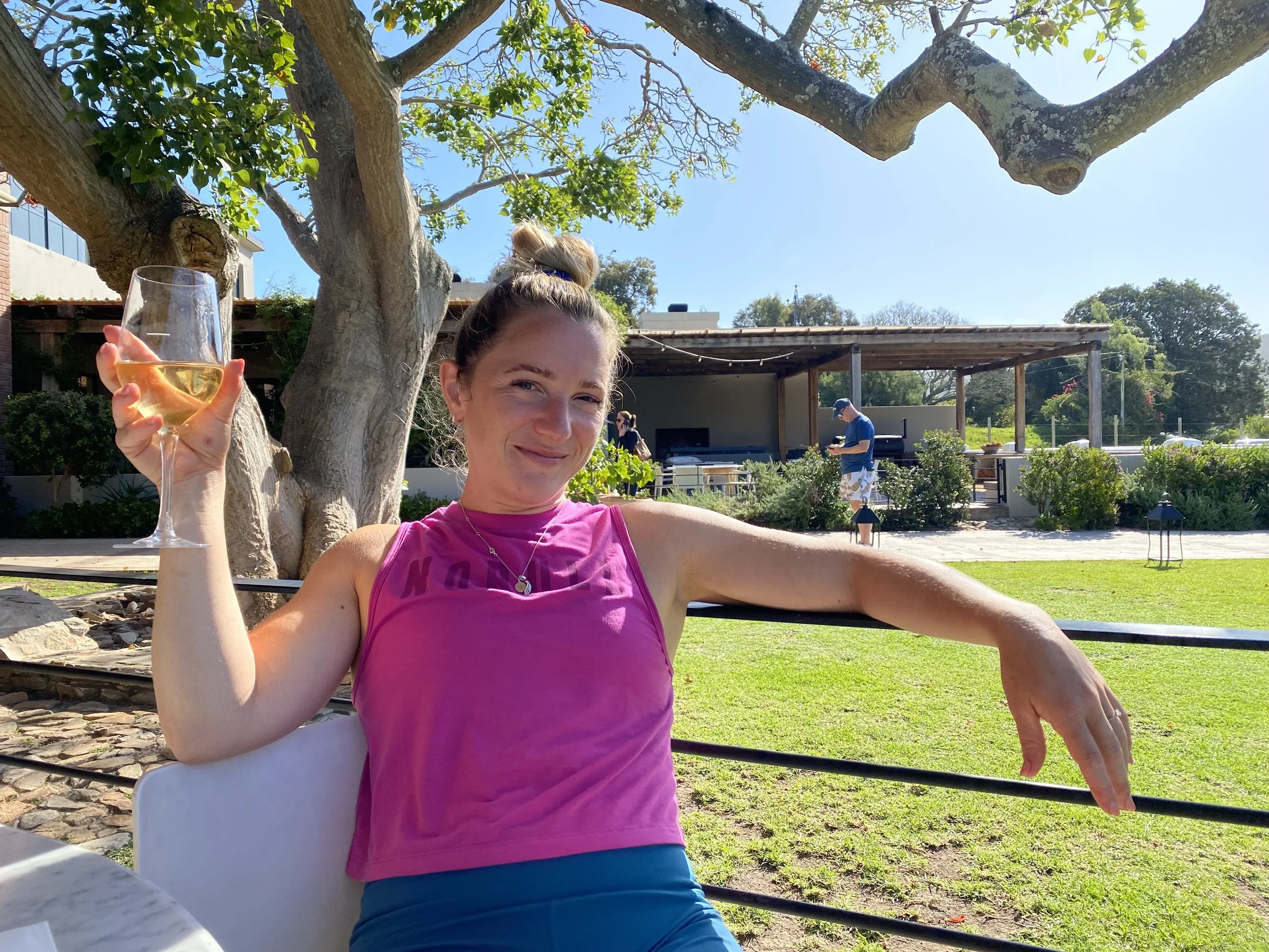 A woman sitting outdoors at a garden party with a glass of white wine, smiling and relaxing under a large tree, with a blue sky and other people in the background.