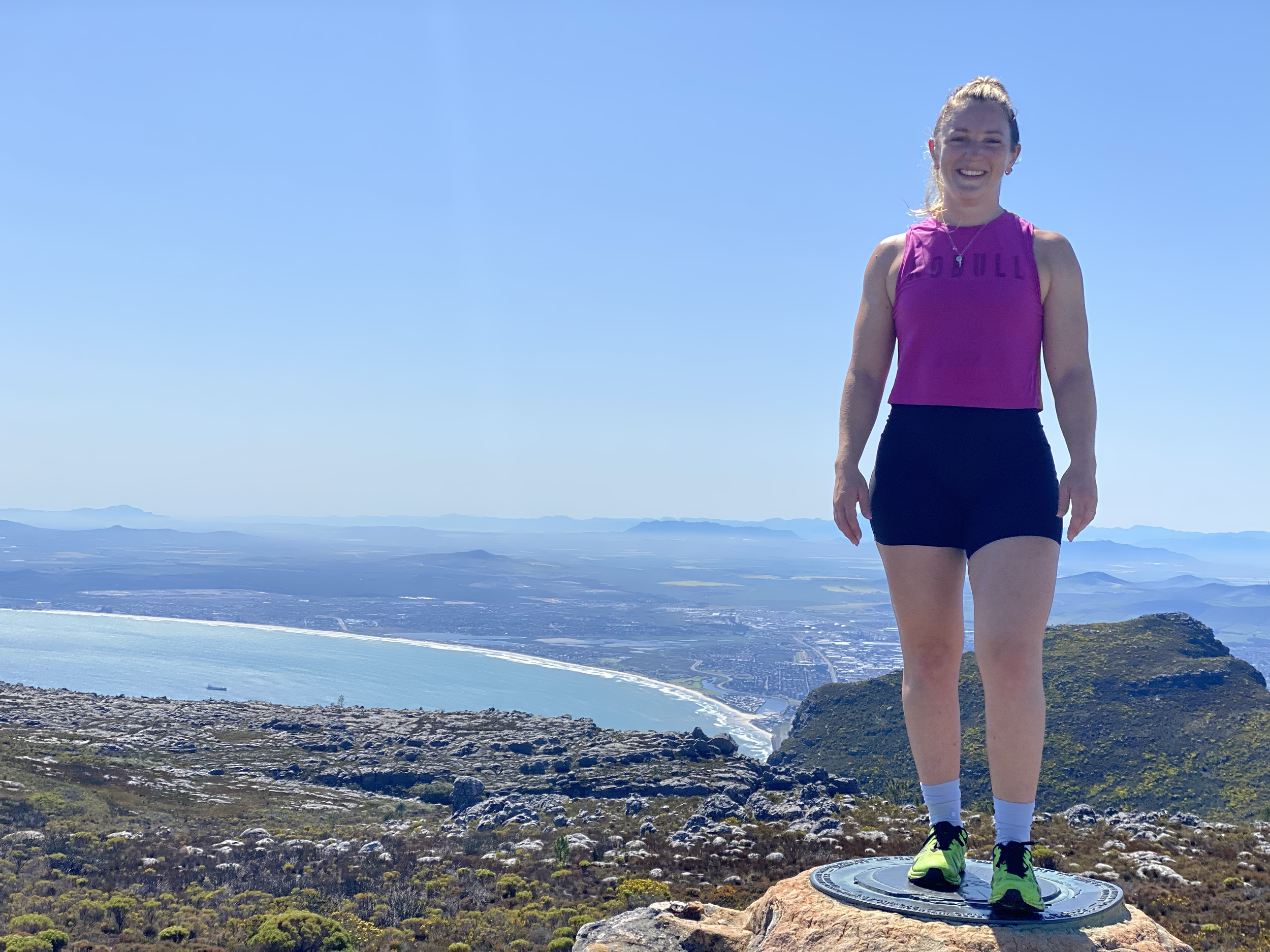 A woman in athletic clothing smiling while standing on a circular platform on a mountain, with a scenic landscape of water, fields, and mountains in the background under a clear blue sky.