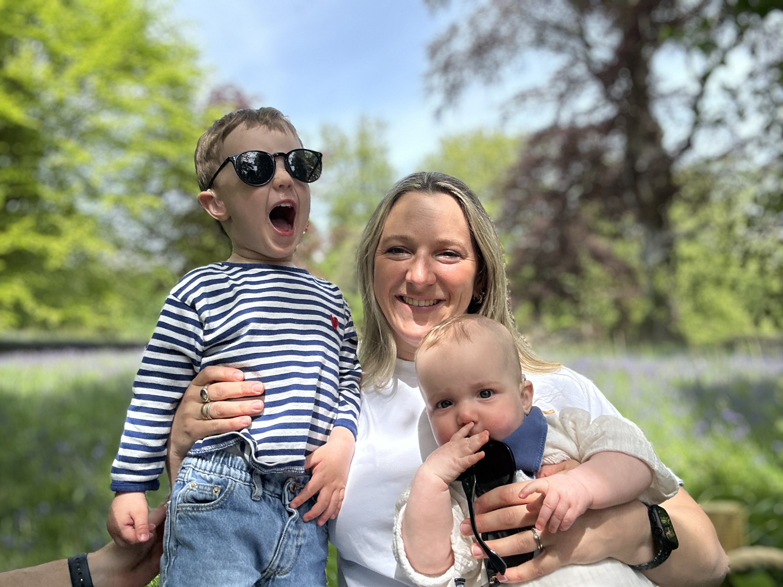A woman with long blonde hair smiling while holding a young boy with sunglasses and a striped shirt, and a toddler with blond hair and a white jacket, in an outdoor park with green trees and blue sky in the background.