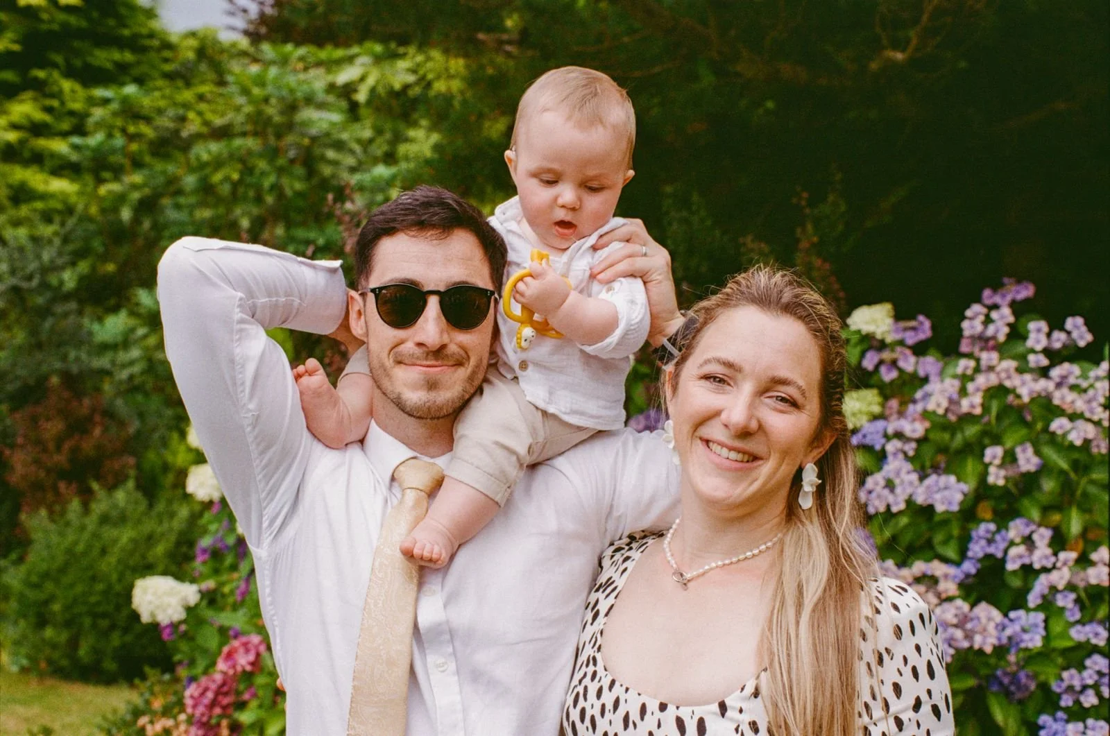 A smiling family of three outdoors in front of blooming flowers. The man is wearing sunglasses and a collared shirt, holding a baby on his shoulder. The woman is smiling at the camera with long blonde hair and polka dot dress.