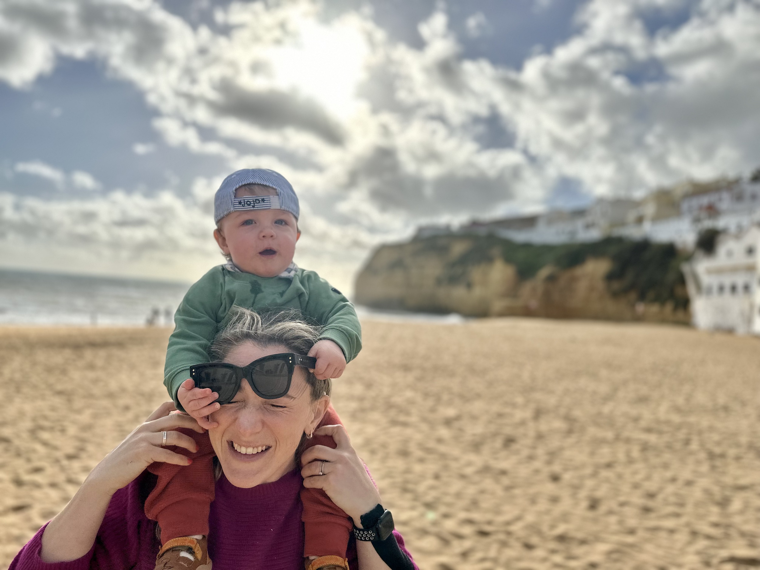 A woman smiling and holding sunglasses on her forehead, carrying a young boy on her shoulders at a sandy beach with cliffs and buildings in the background on a partly cloudy day.