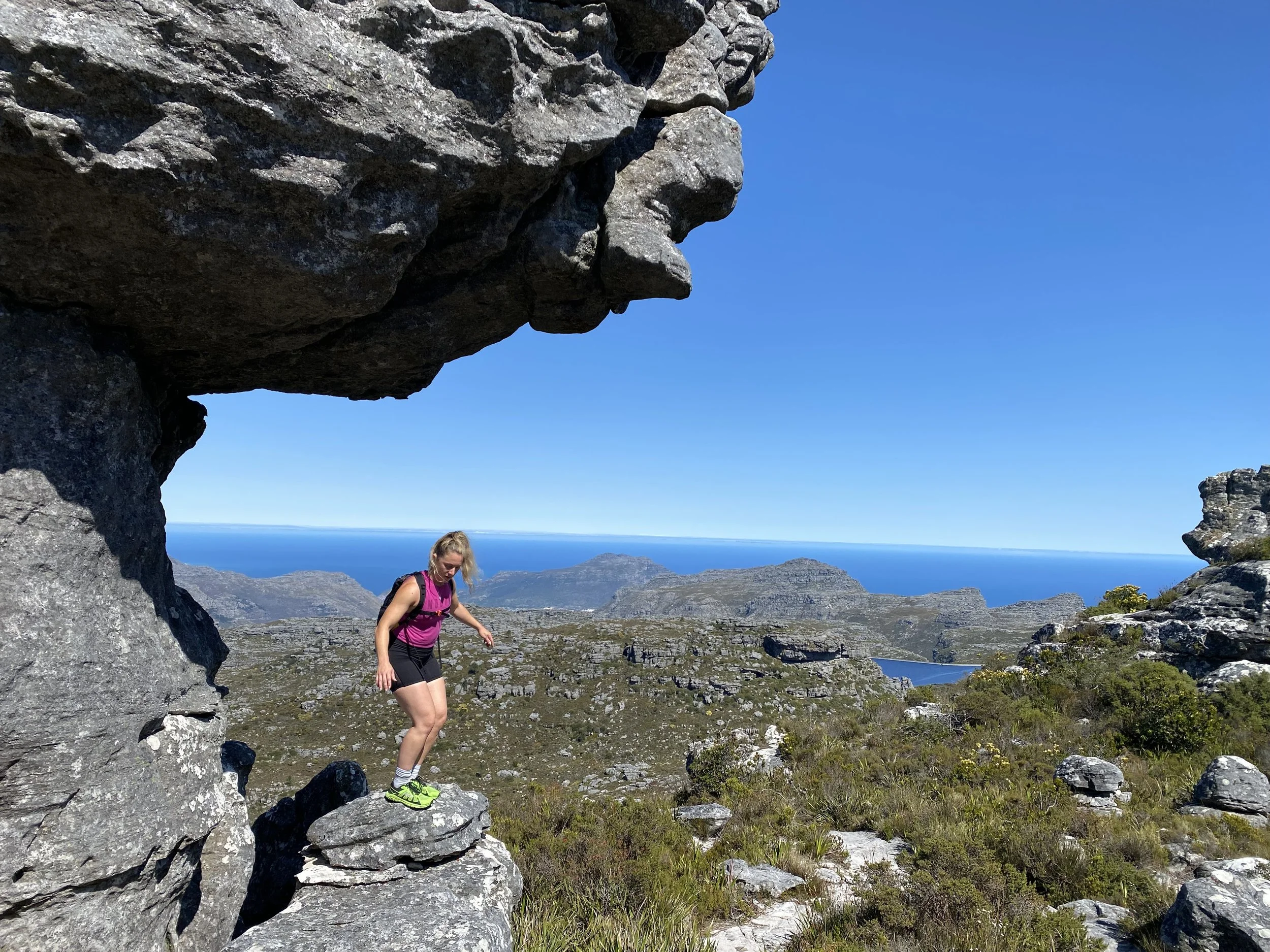 A woman hiking in a rocky mountain landscape with a clear blue sky and distant ocean in the background.