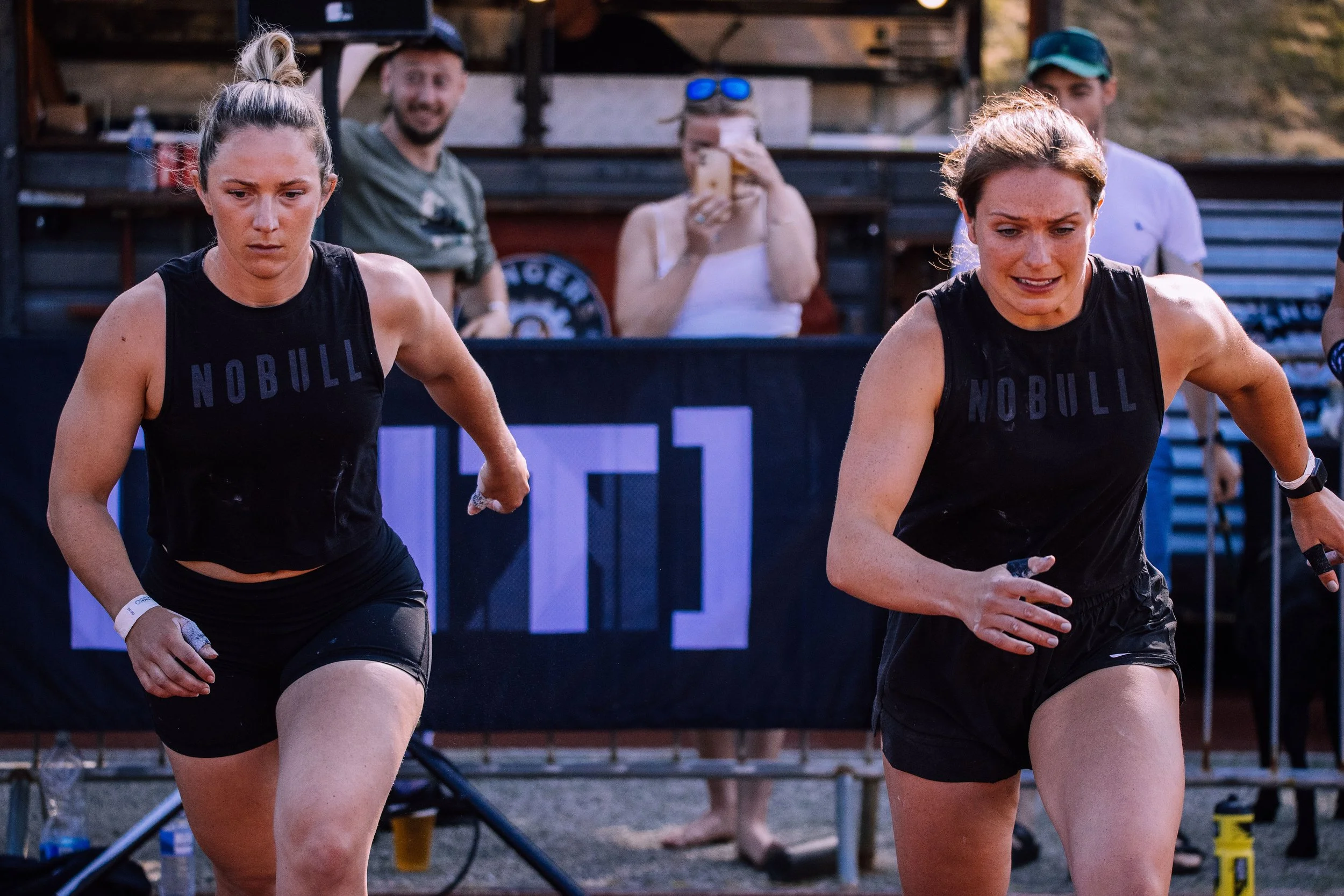 Two female athletes in black athletic attire running in a race at an outdoor event, with spectators and equipment visible in the background.