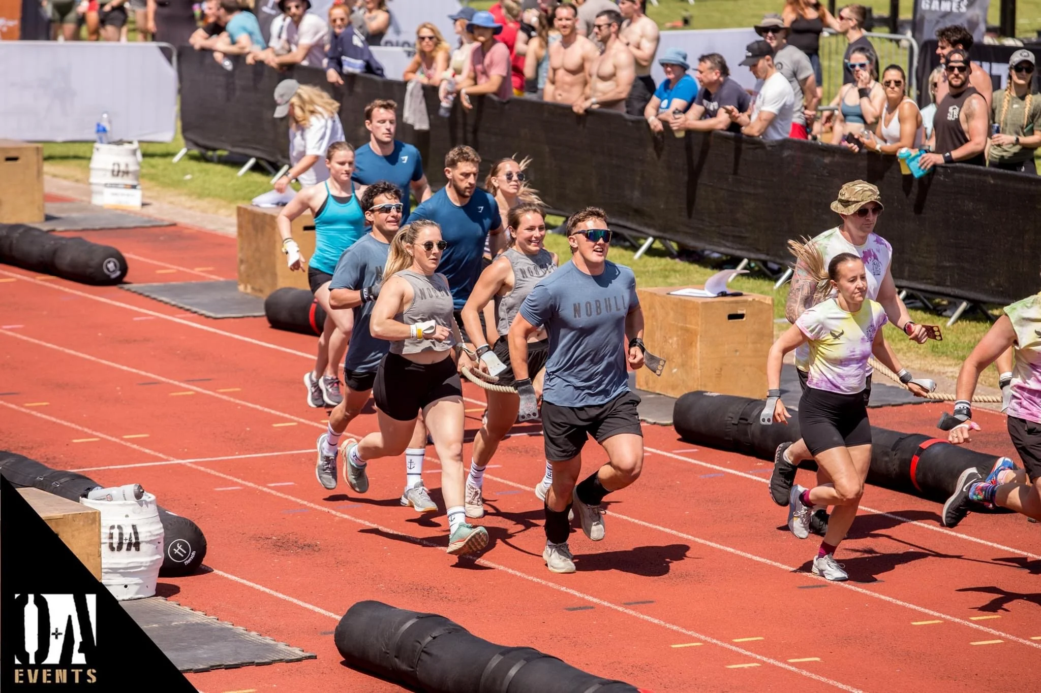 Group of people participating in an outdoor fitness or military-style obstacle course event on a running track with black barriers and tires. Spectators watch behind a black fence.
