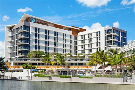 Modern multi-story hotel with glass balconies and palm trees in front, situated by the water under a blue sky.
