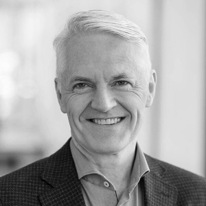 Black and white headshot of a smiling professional with short, light-colored hair, wearing a suit jacket and dress shirt.