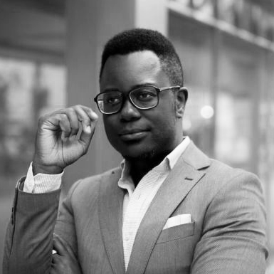 A professional in a suit adjusts their glasses while looking at the camera, standing outdoors with a blurred city background.