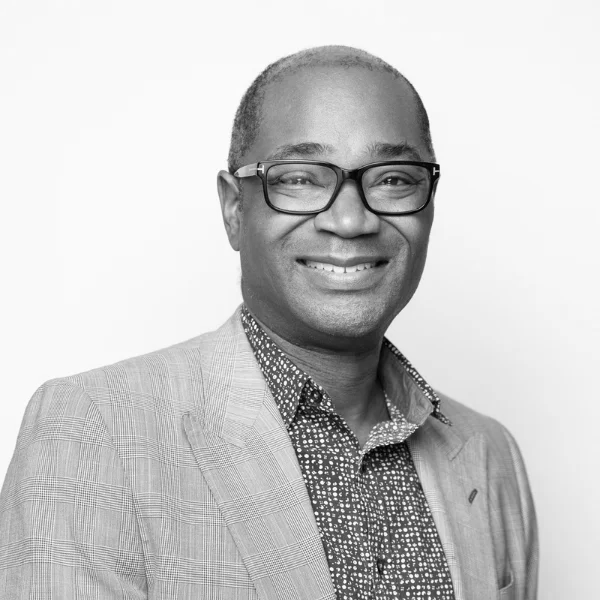 A black and white portrait of a smiling professional wearing glasses, a checkered shirt, and a blazer.