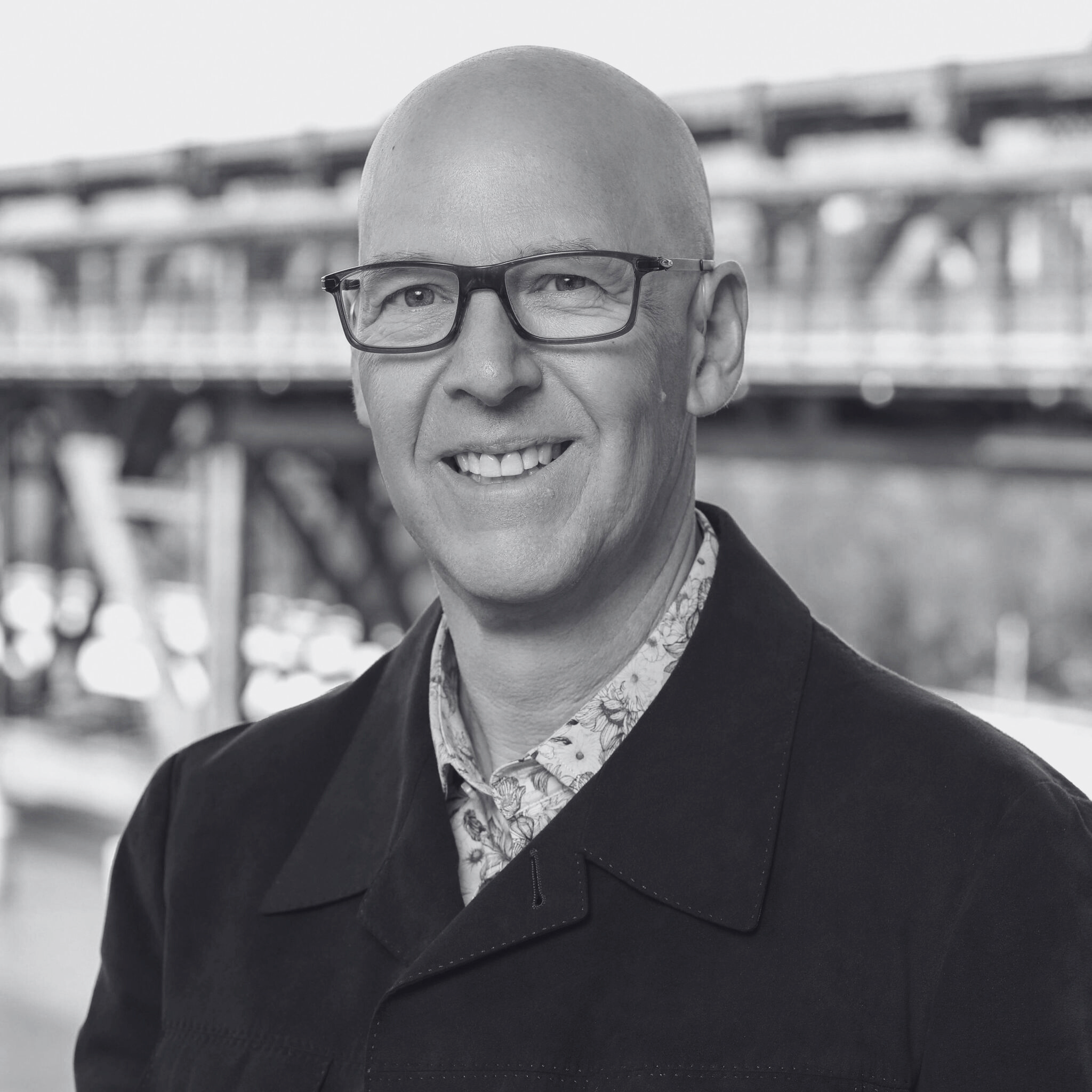 Close-up of a professional with a bald head and glasses, smiling, wearing a patterned shirt under a jacket, with a blurred bridge or industrial structure in the background.