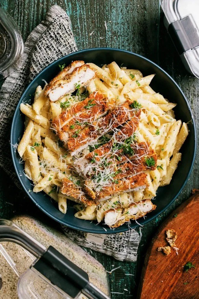 A bowl of penne pasta topped with breaded and fried chicken cutlet, grated cheese, and garnished with chopped parsley.