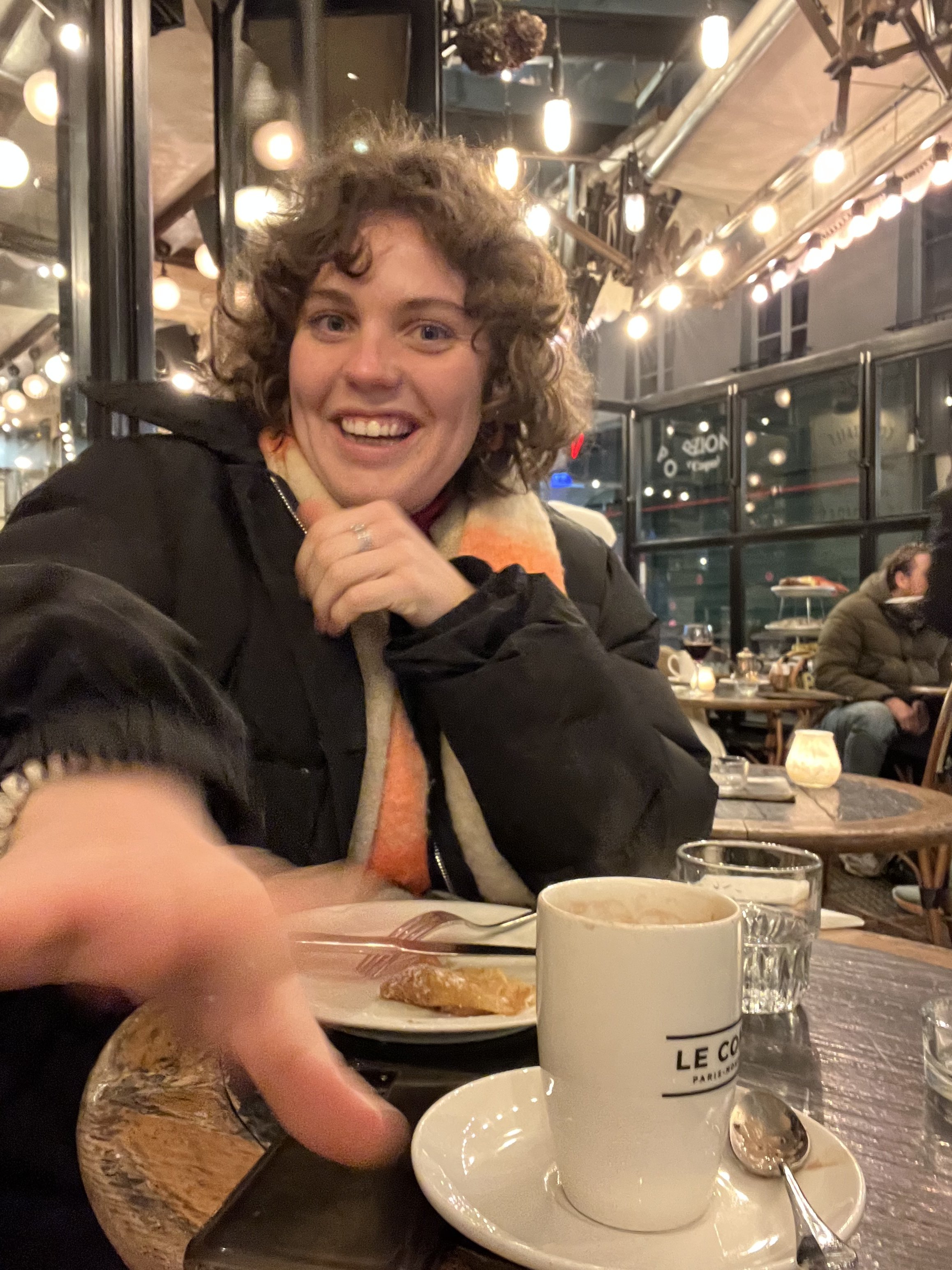 Une femme souriante dans un restaurant, devant une tasse de café, avec d'autres personnes en arrière-plan, et des lumières chaudes suspendues au plafond.