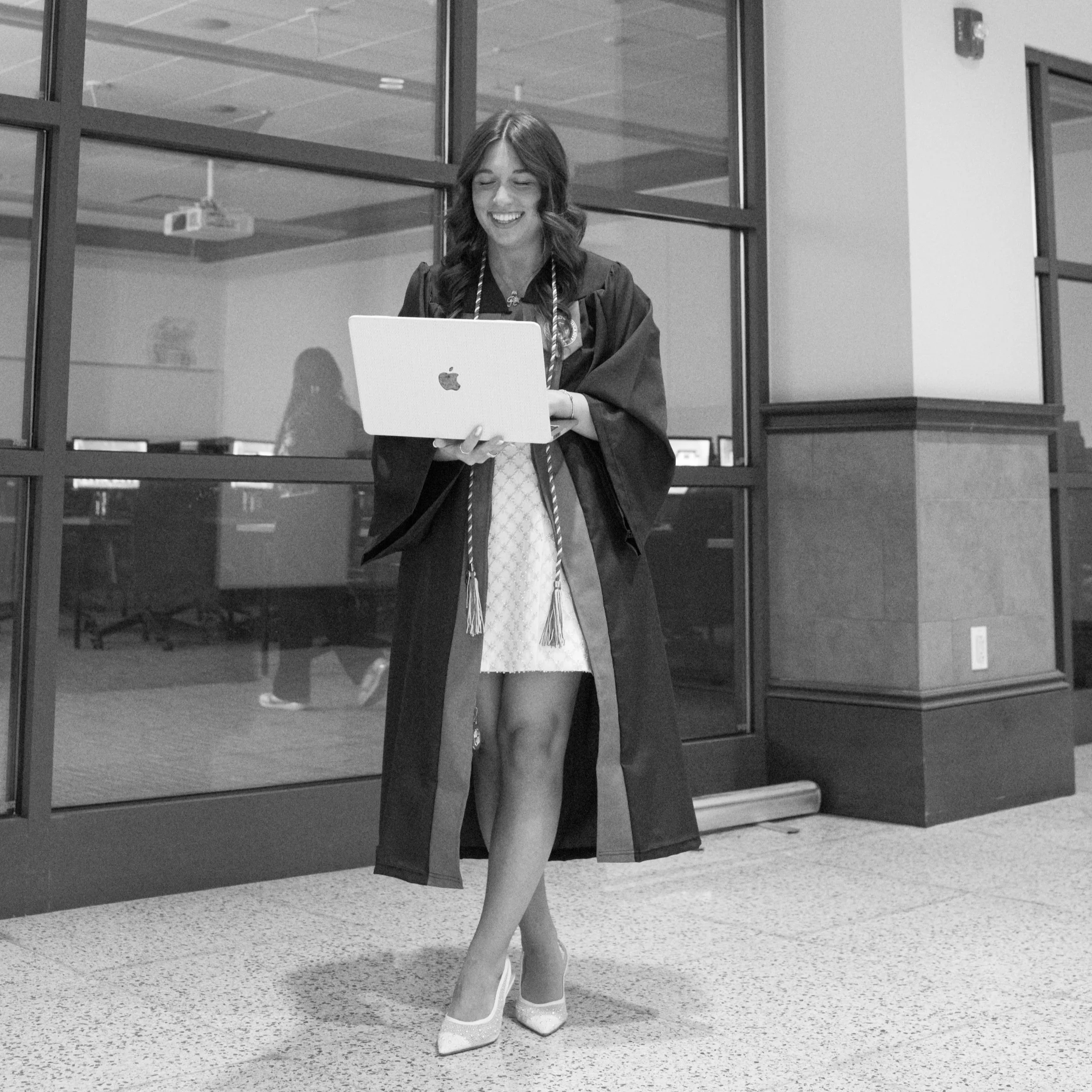 Woman in cap and gown looking at a laptop, standing in an office or school hallway.