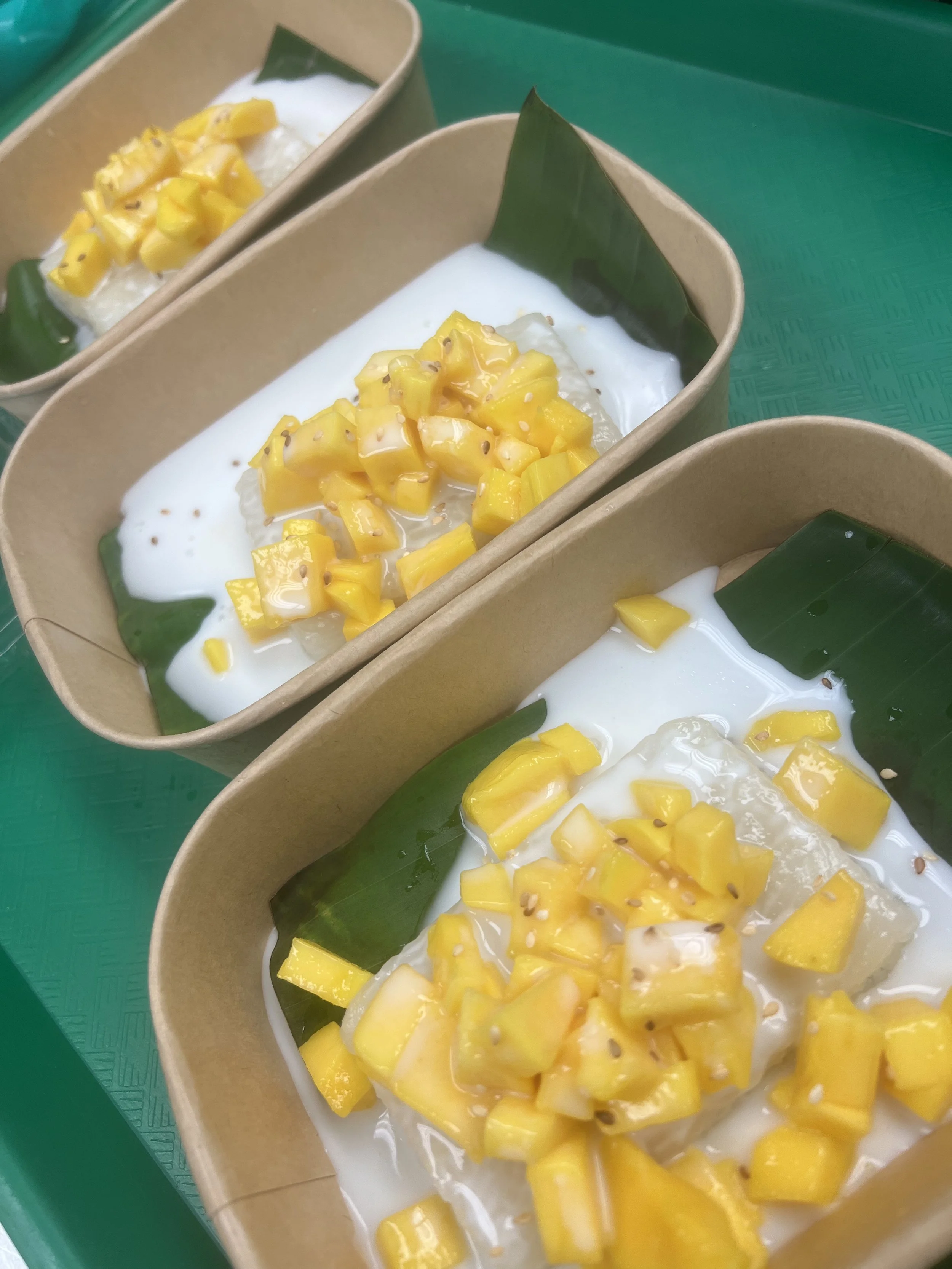 Close-up of three takeaway containers filled with coconut milk topped with diced yellow mango pieces, placed on a green surface.