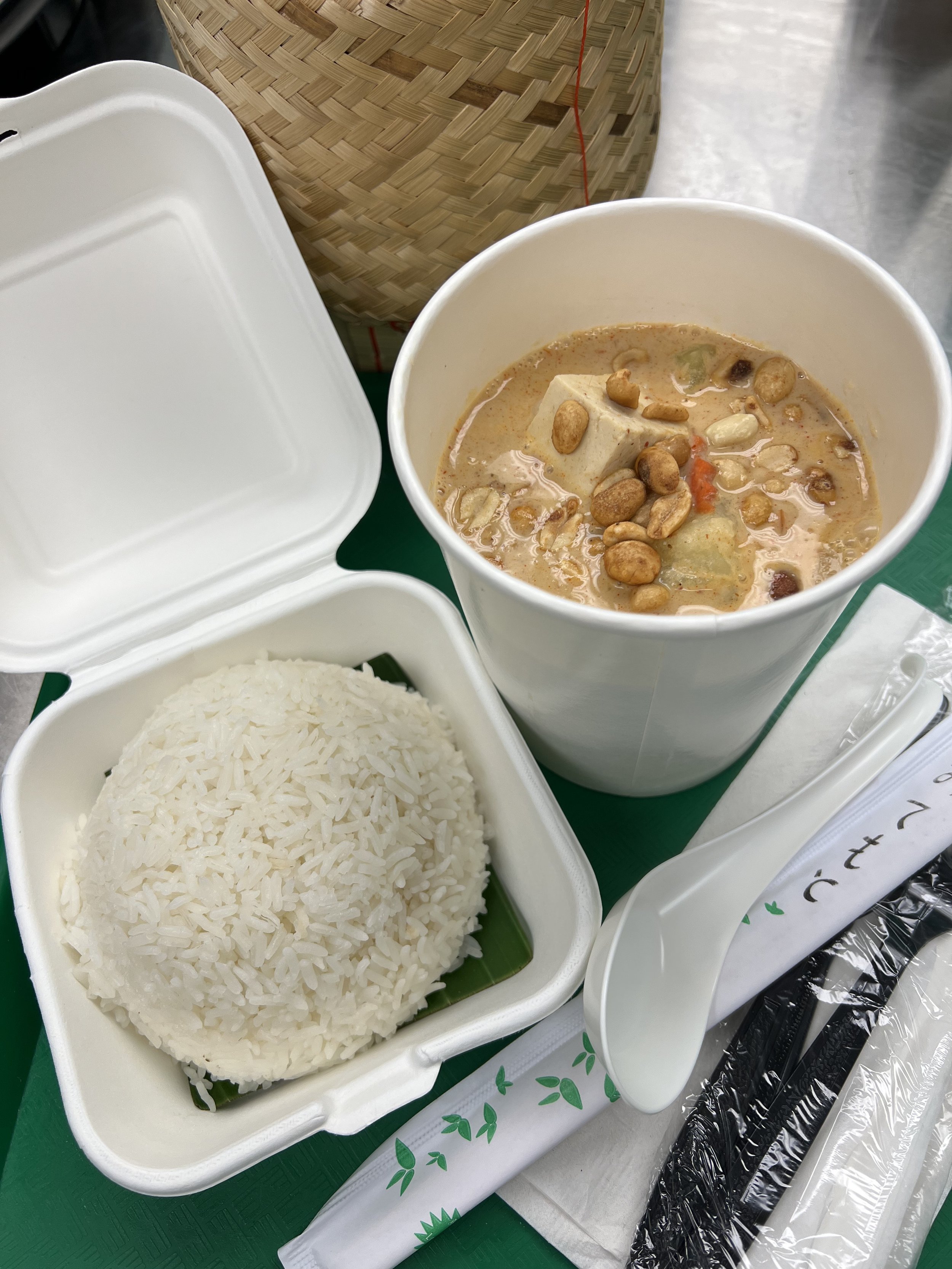 A tray of steaming white rice in a white takeout container, a cup of curry with tofu and vegetables topped with peanuts, and part of a bamboo container and utensils on a table.