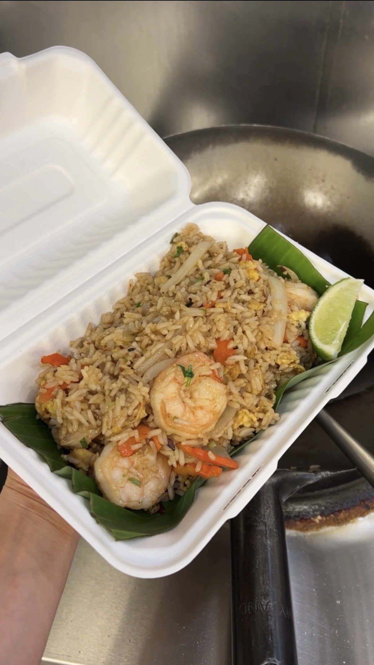 A takeout container of shrimp fried rice garnished with lime wedge and green leaves, held above a stove.