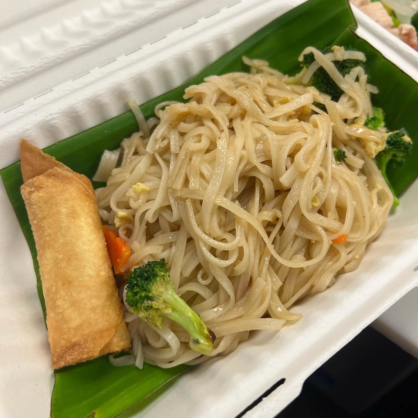 Takeout container with stir-fried lo mein noodles, broccoli, a spring roll, and a small piece of carrots on a green leaf liner.