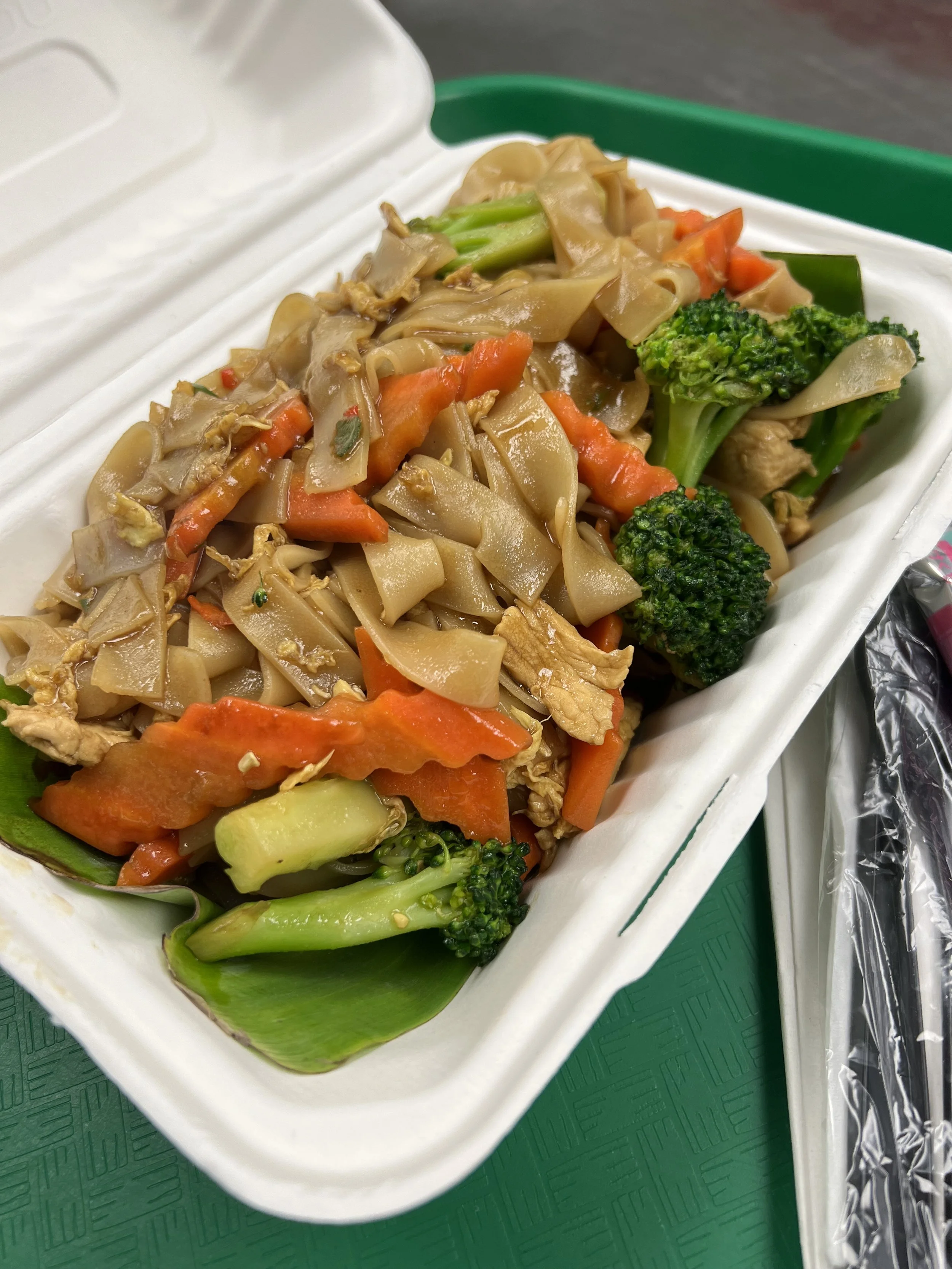 To-go container filled with stir-fried noodles, broccoli, carrots, and other vegetables on a green tray with a plastic fork wrapped in foil.