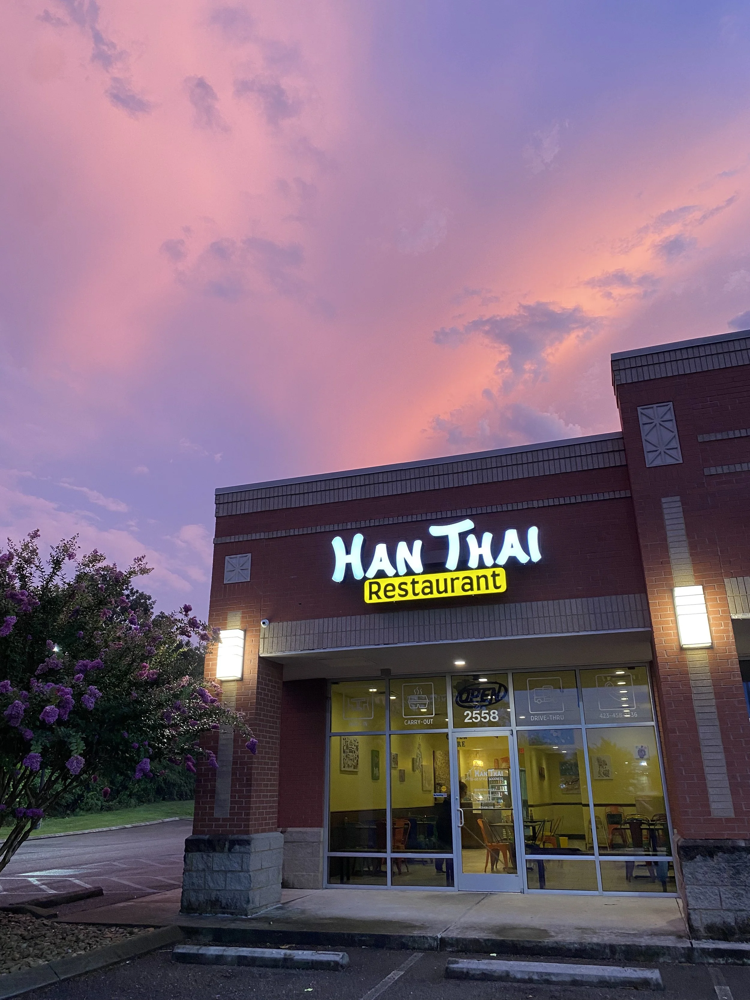 Exterior view of Han Thai restaurant at dusk, showing a brick building with a brightly lit sign, large front windows, and pink and purple clouds in the sky, with flowering bushes in the foreground.