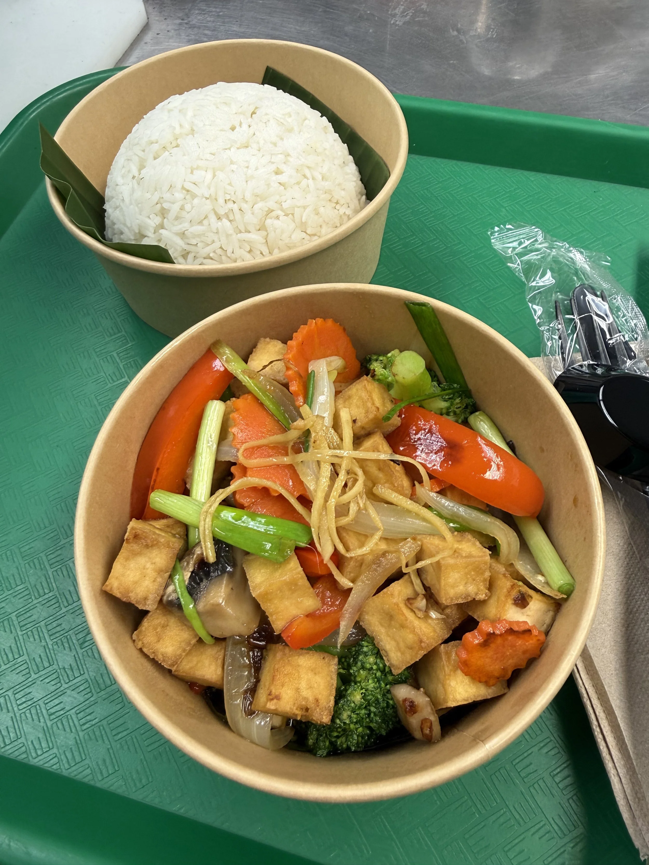 Disposable food container with rice and a separate container with stir-fried vegetables and tofu, placed on a green tray.