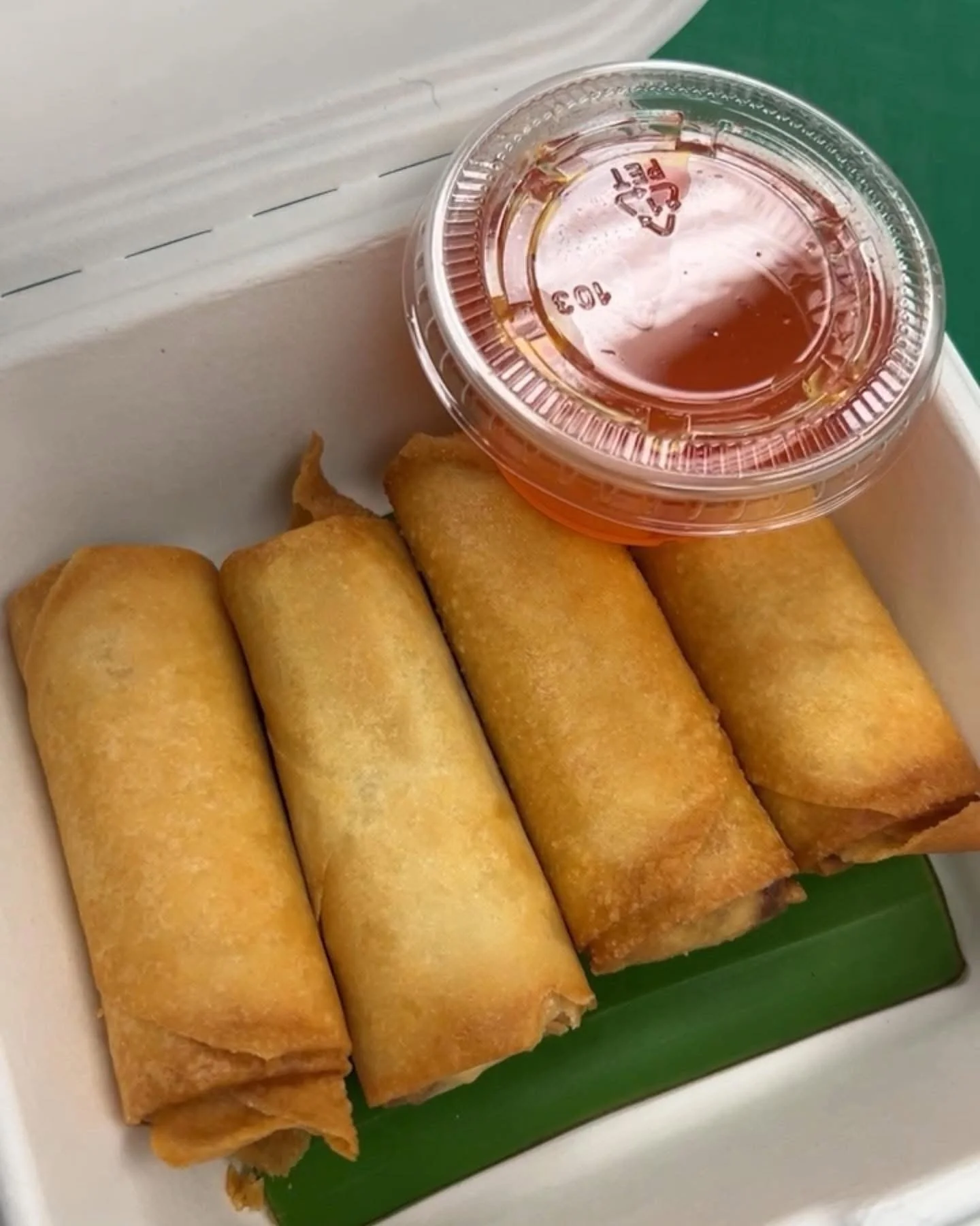 Four fried spring rolls in a white takeout box with a green insert, accompanied by a small plastic container of sweet and sour sauce on the side.