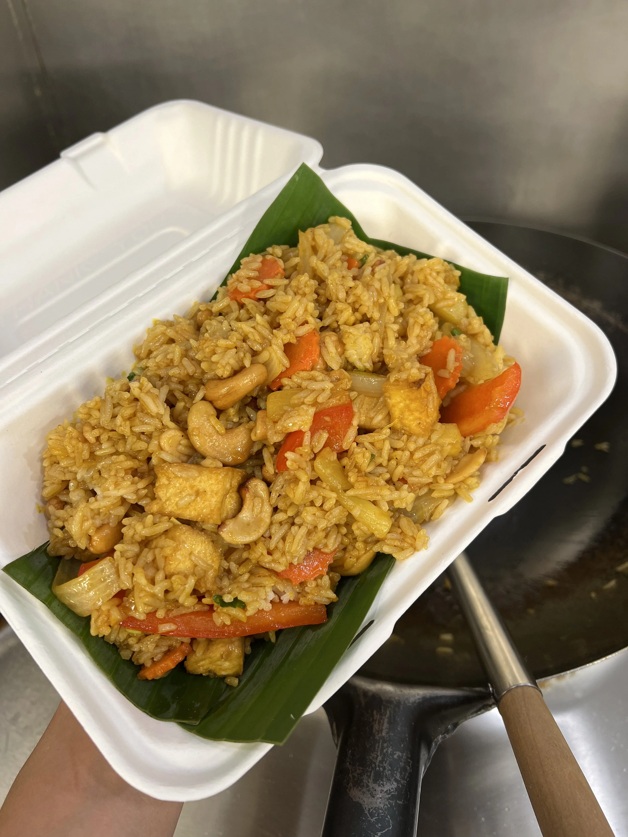 A takeout container of fried rice with vegetables, placed on a green banana leaf, being held over a wok stove.