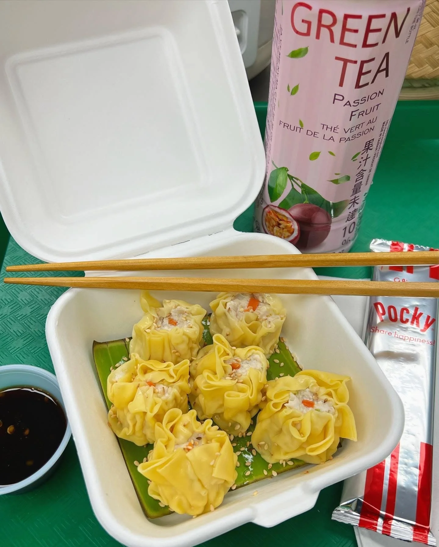 Steamed dumplings in a white takeout container, soy sauce in a small dish, a packet of Pocky chocolate snack, a can of Green Tea Passion Fruit drink, and a pair of wooden chopsticks on top of the container.