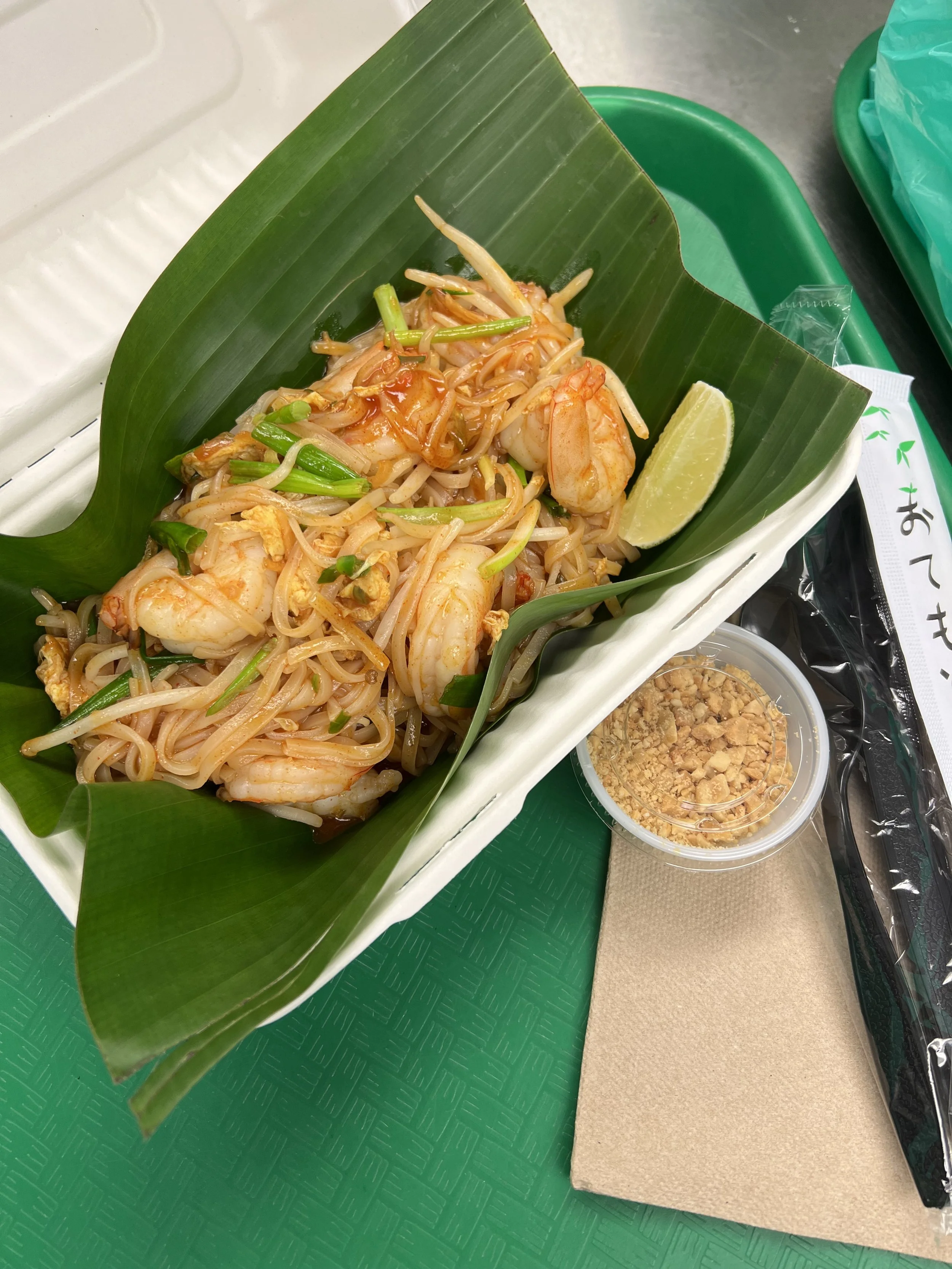 A plate of Thai Pad Thai noodles with shrimp garnished with bean sprouts, green onions, and a wedge of lime, served on a green leaf on a white tray.